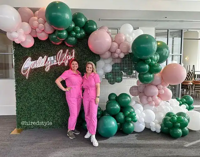 Two women in matching pink jumpsuits stand in front of a stunning balloon installation and a neon sign reading "Giddy Up!" on a faux grass wall, showcasing the creativity of a Los Angeles balloon artist.