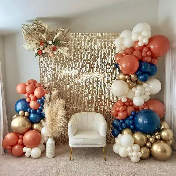 A white chair sits in front of a gold sequin backdrop, decorated with elaborate balloon installations in white, gold, blue, and peach by a Los Angeles balloon artist, accented with dried floral arrangements.