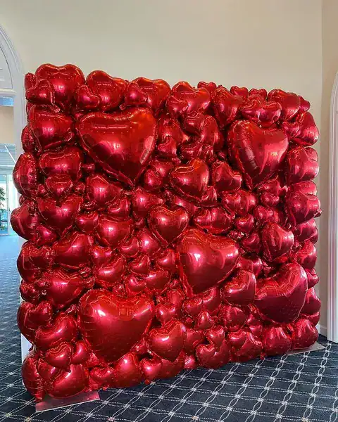 A wall display created by a balloon artist features numerous shiny red heart-shaped balloons arranged closely together inside a room with arched doorways.