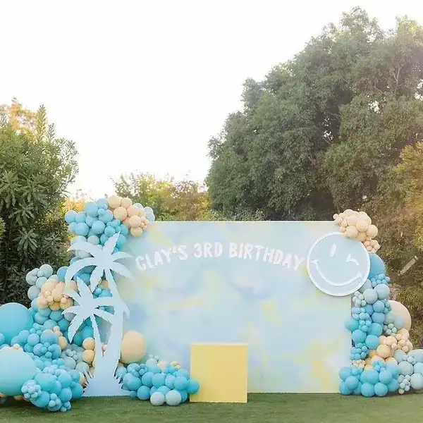 Outdoor birthday backdrop decorated with pastel blue, yellow, and beige balloons by a skilled balloon artist, featuring a palm tree cutout and a large smiley face; text reads "Clay's 3rd Birthday.”.