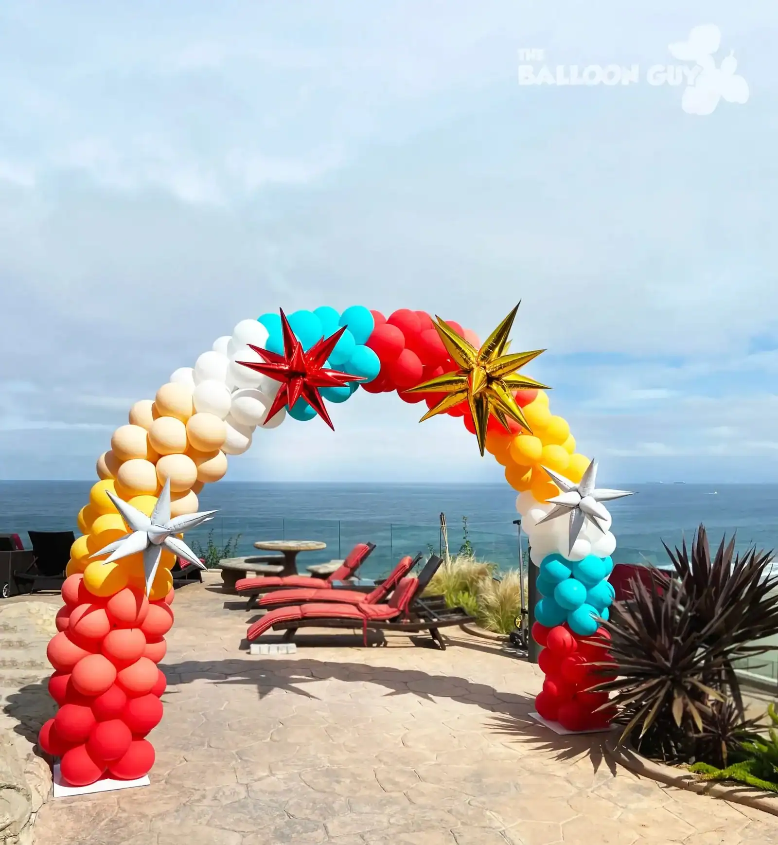 A balloon arch in red, white, yellow, and blue with metallic star decorations stands on a patio overlooking the ocean in Los Angeles, perfect for corporate event balloon decor amidst lounge chairs and plants.