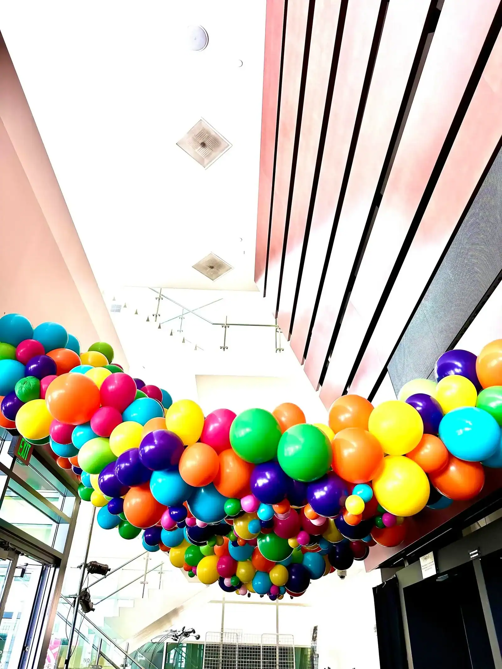 A colorful balloon garland, crafted by a skilled balloon artist, is suspended across the ceiling of a modern indoor space with large windows and a staircase in the background.
