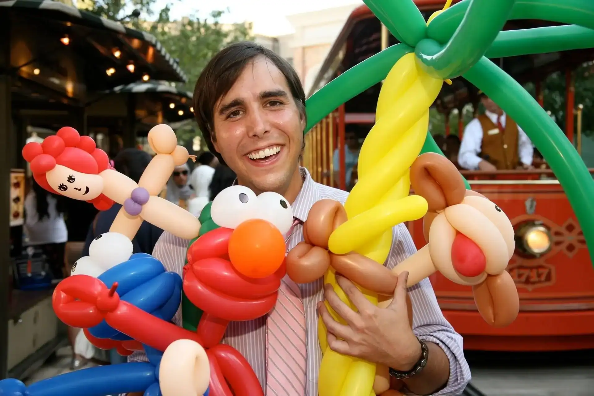 A man smiling and holding several colorful balloon animals, including Elmo and a palm tree, at an outdoor corporate event in Los Angeles.