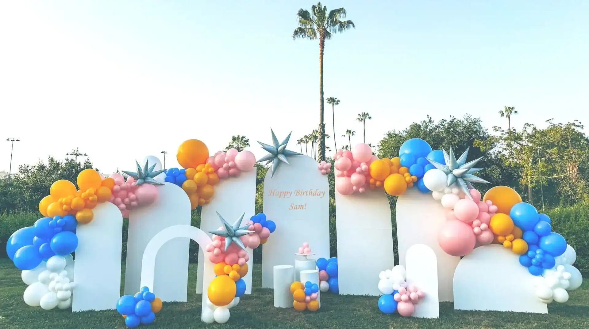 Outdoor birthday party setup in Los Angeles with stunning balloon decor—white arches, colorful balloon clusters in blue, pink, orange, and silver, and a "Happy Birthday Sani" sign. Palm trees provide a perfect backdrop for this festive balloon installation.