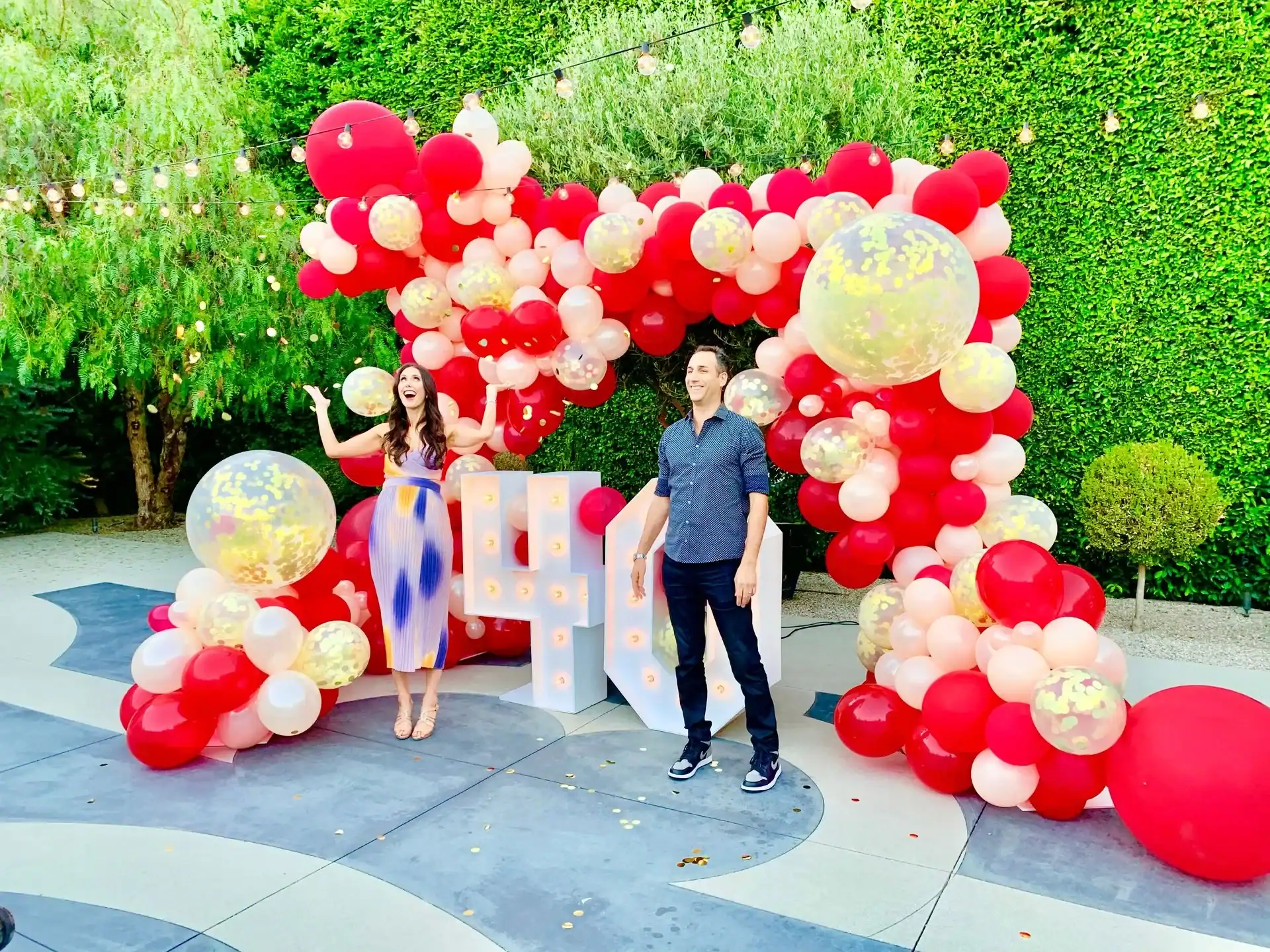 Two people stand in front of a stunning balloon decor arch with large light-up "40" numbers, outdoors among greenery and string lights.