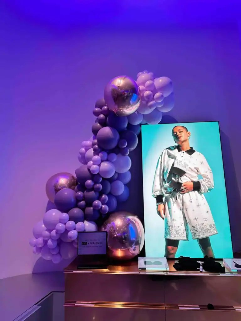 A digital display shows a person in white clothing beside a decorative arrangement of purple and silver balloons, showcasing balloon installations under blue-purple lighting at a corporate event in Los Angeles.