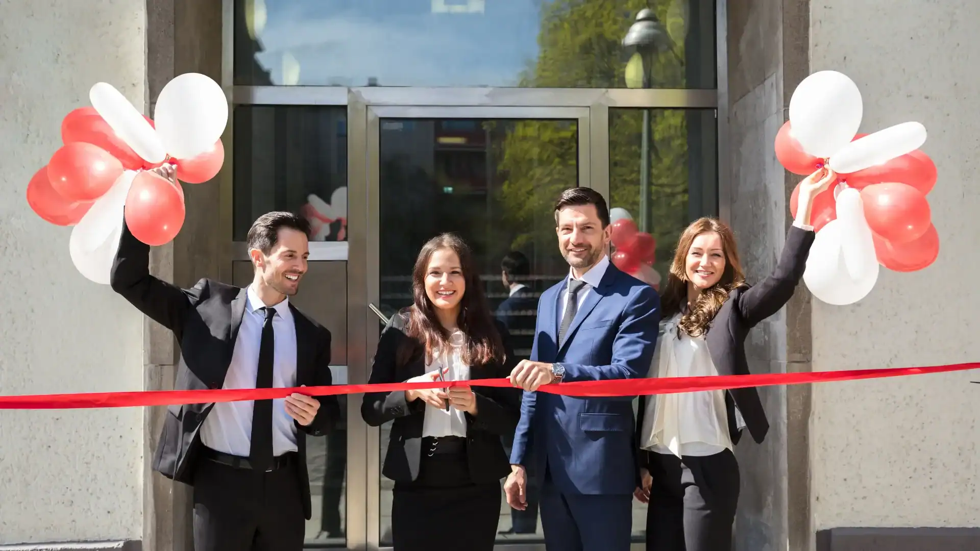 Four people in business attire stand in front of a Los Angeles building, holding balloons and cutting a red ribbon at a ribbon-cutting ceremony featuring vibrant balloon installations.