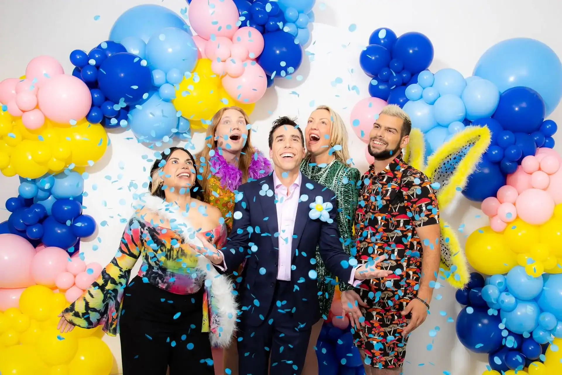 Five people stand smiling under colorful balloons created by a skilled balloon artist as blue confetti falls around them, posing for a celebratory group photo at a lively corporate event against a white background.