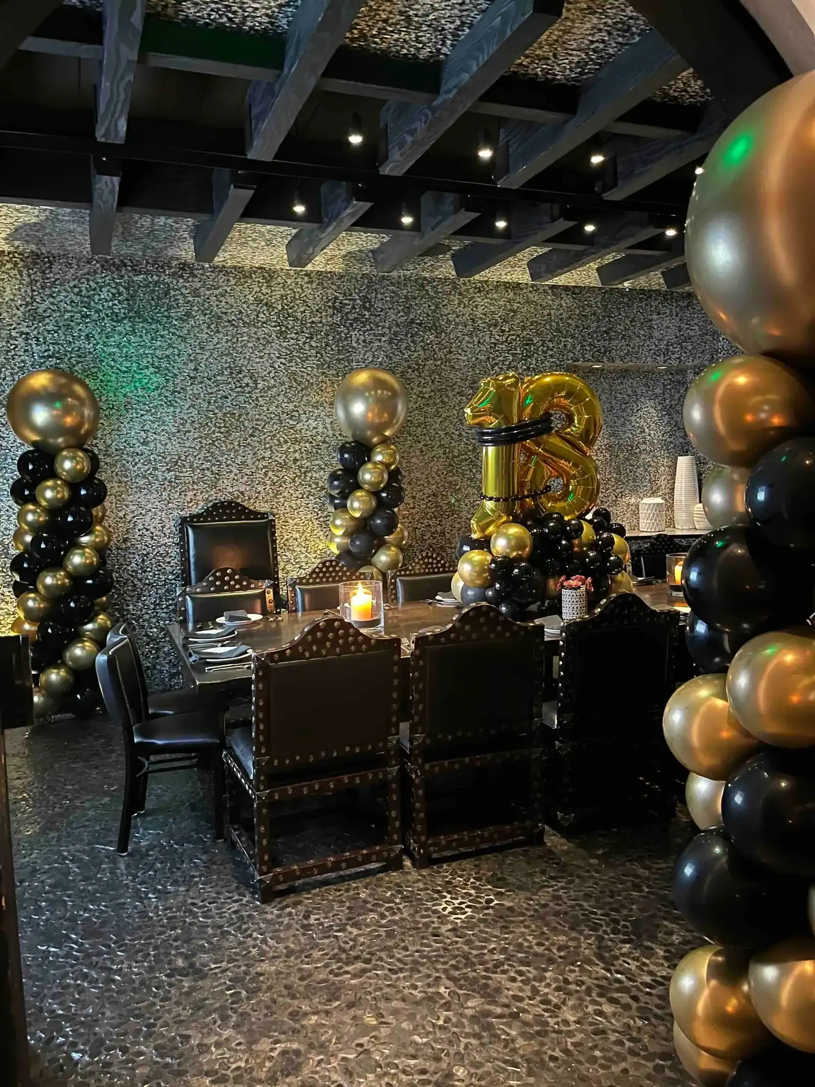 A dining area decorated with black and gold balloons by a balloon artist, featuring a large "23" balloon arrangement, set for a celebration or corporate event with six chairs around the table.