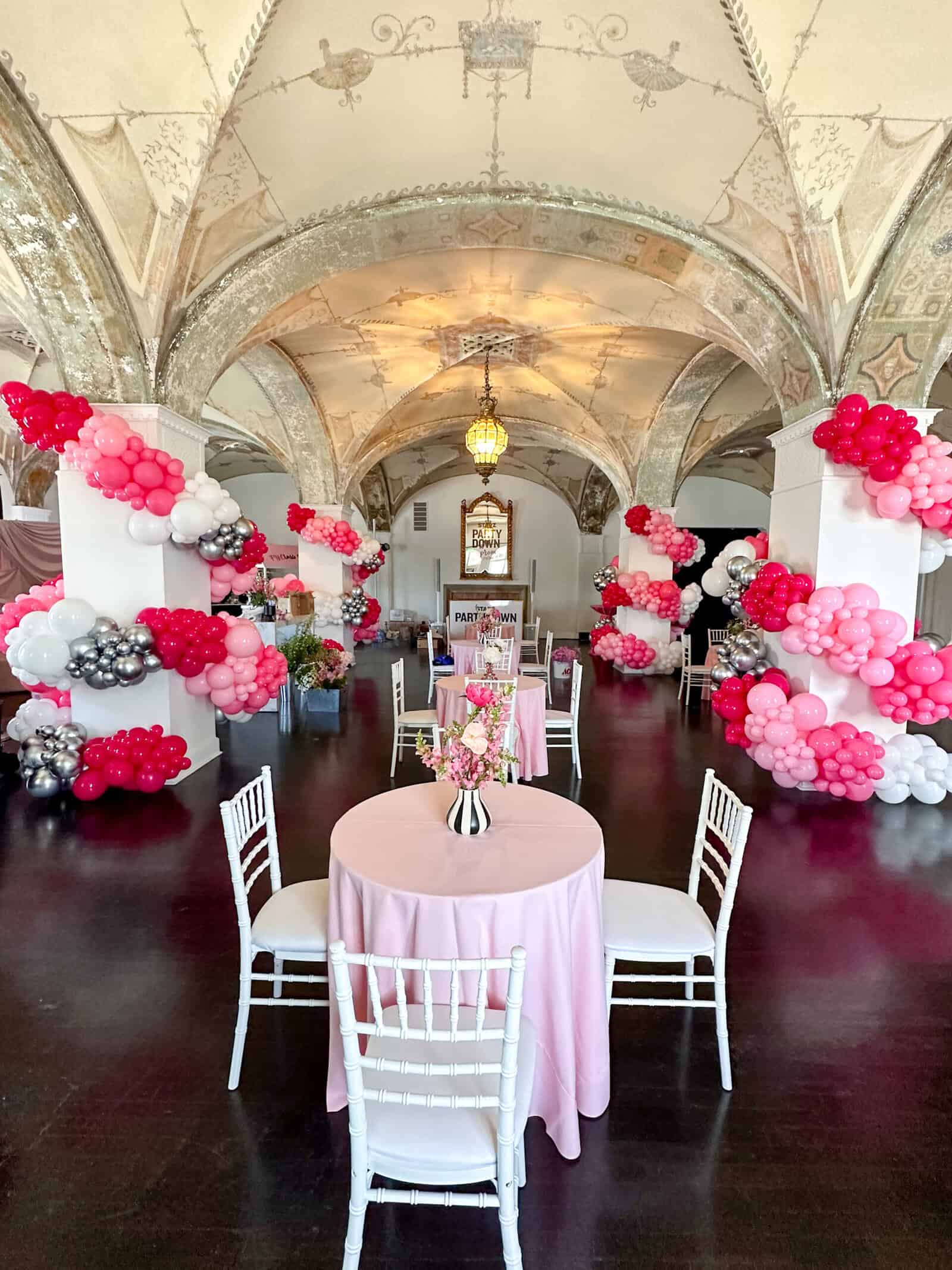A decorated event hall in Los Angeles with arched ceilings, pink and white balloon arrangements crafted by a skilled balloon artist, round tables with pink tablecloths, and white chairs.