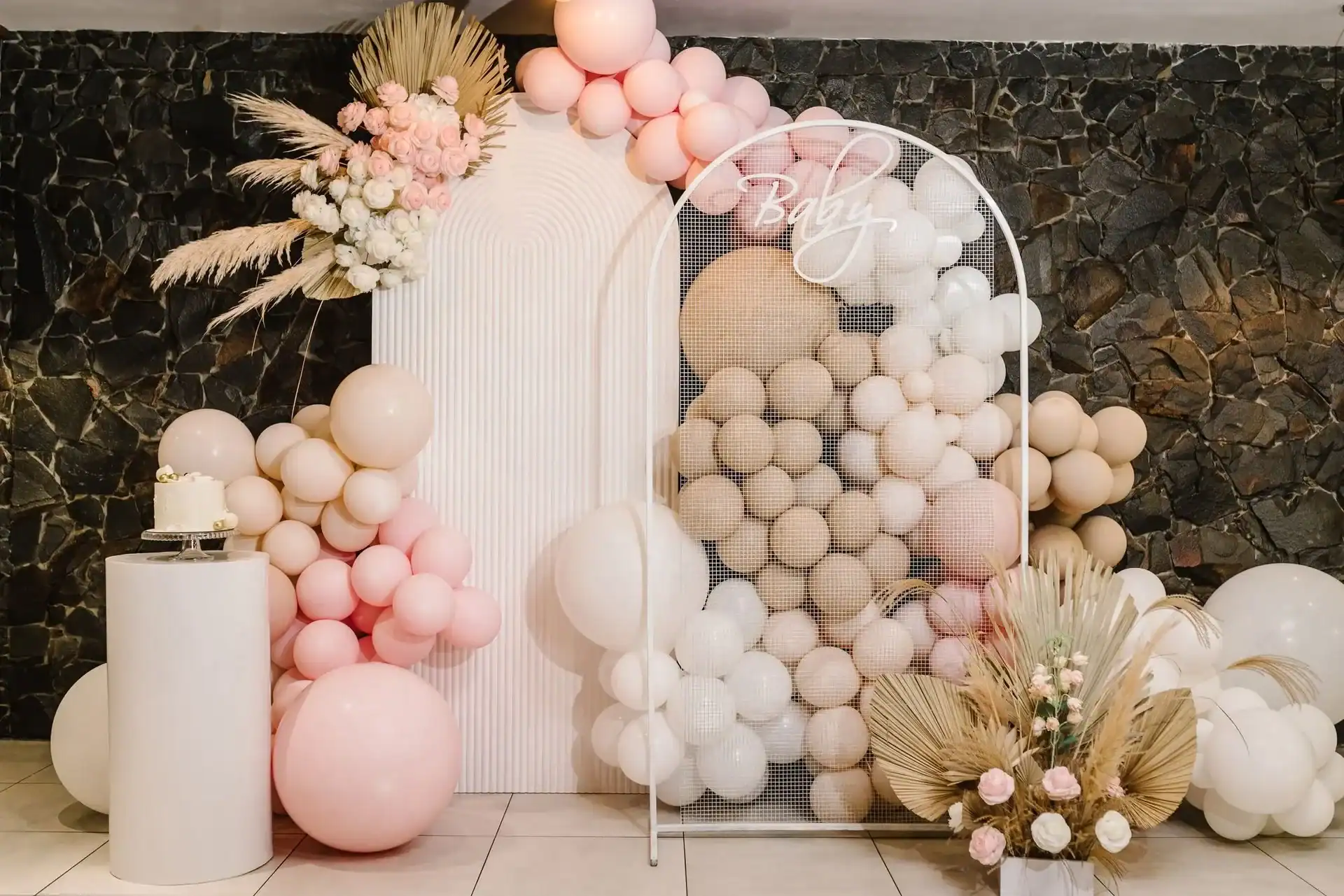 A baby shower display in Los Angeles featuring pink, beige, and white balloons, dried grasses, flowers, and a "Baby" sign on a mesh arch against a dark stone wall.