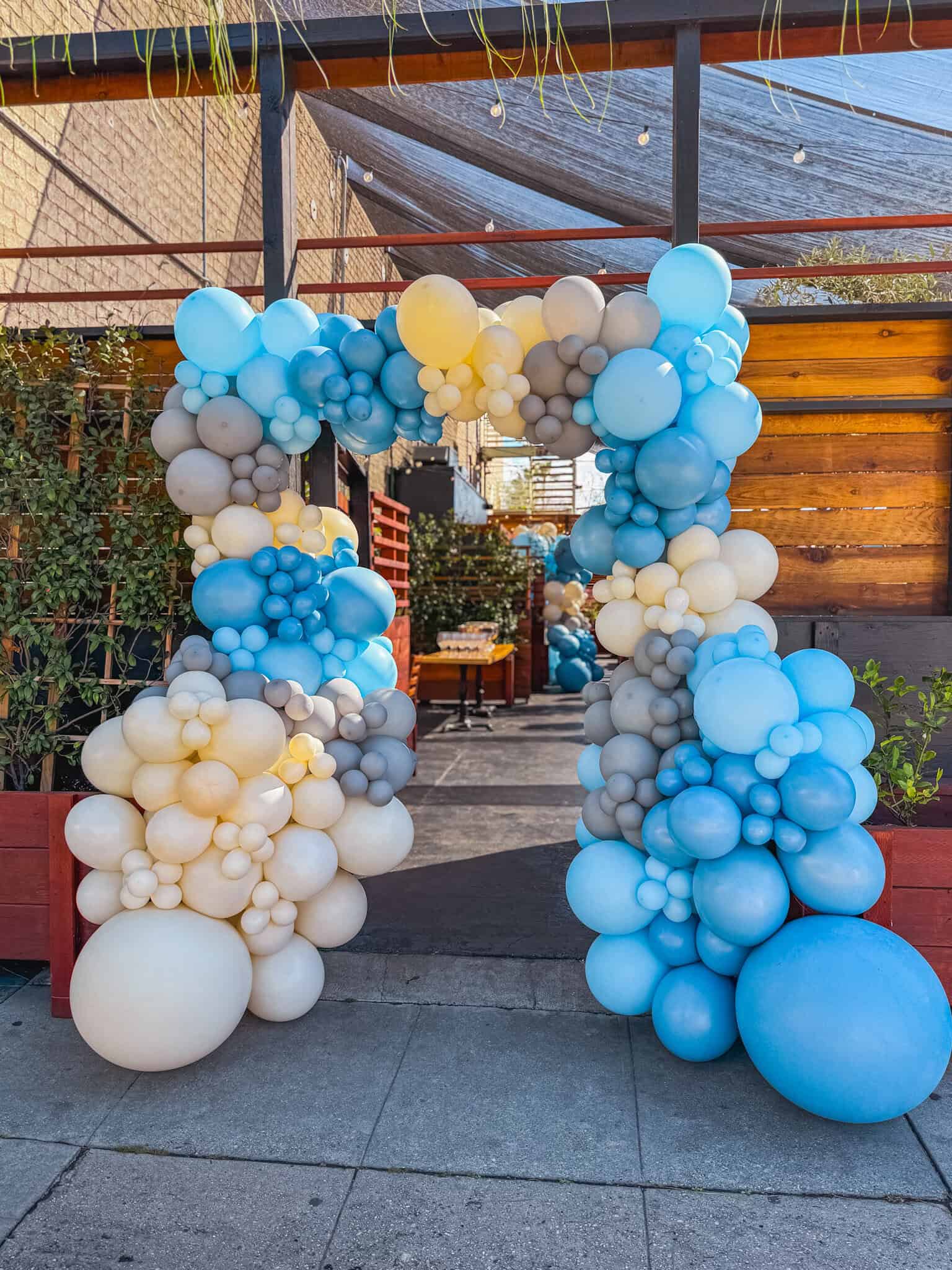 A decorative arch made of blue, white, and gray balloon decor by a skilled balloon artist stands at the entrance to an outdoor patio area with wooden walls and greenery in Los Angeles.
