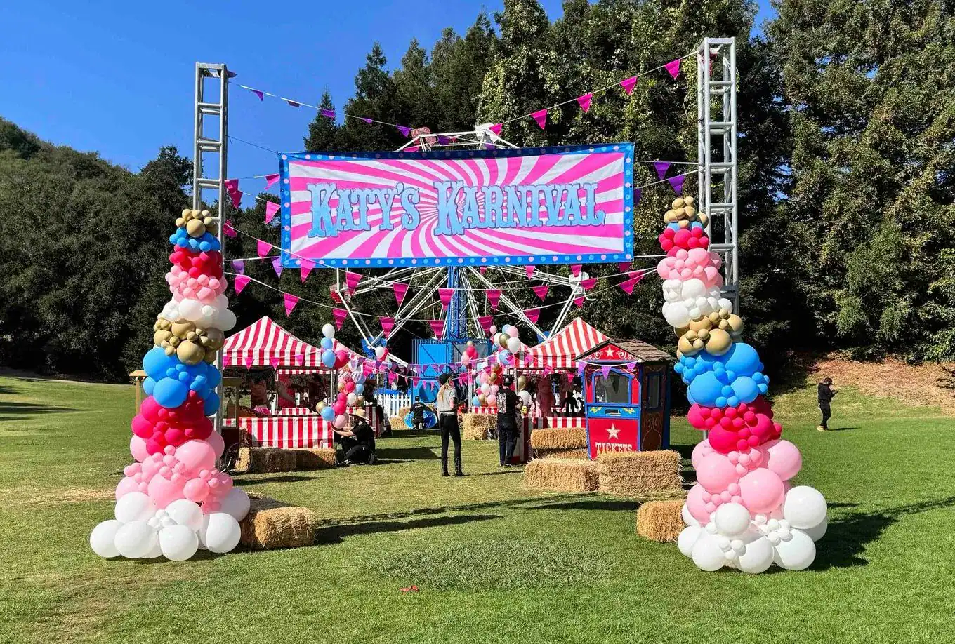 Outdoor carnival setup with striped tents, vibrant balloon installations, hay bales, and a large sign reading "Mary's Carnival" on a grassy field surrounded by trees—perfect for a fun-filled corporate event.
