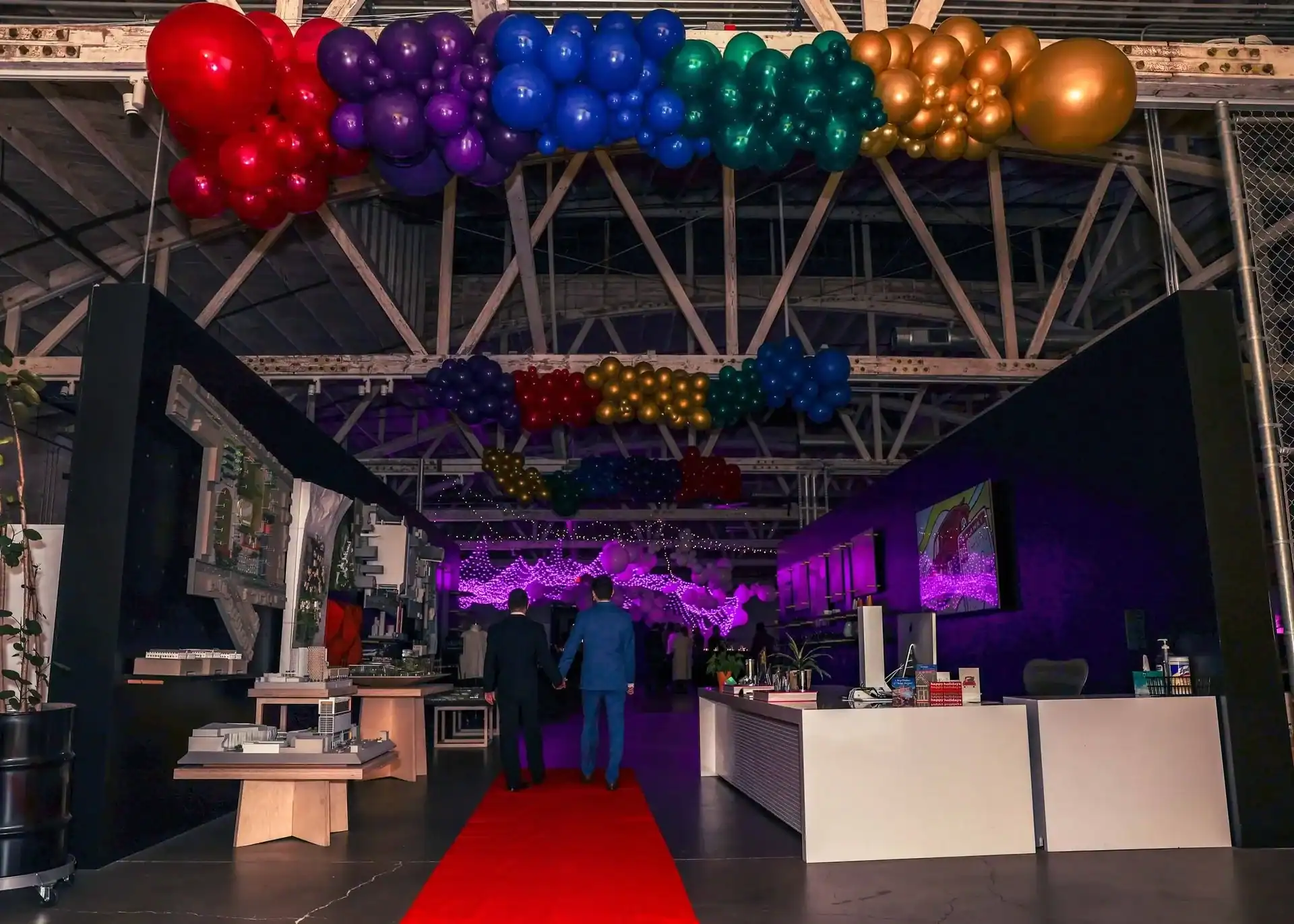 A red carpet leads into an industrial-style Los Angeles event space decorated with colorful balloon installations on the ceiling and purple lighting in the background, with people walking inside.