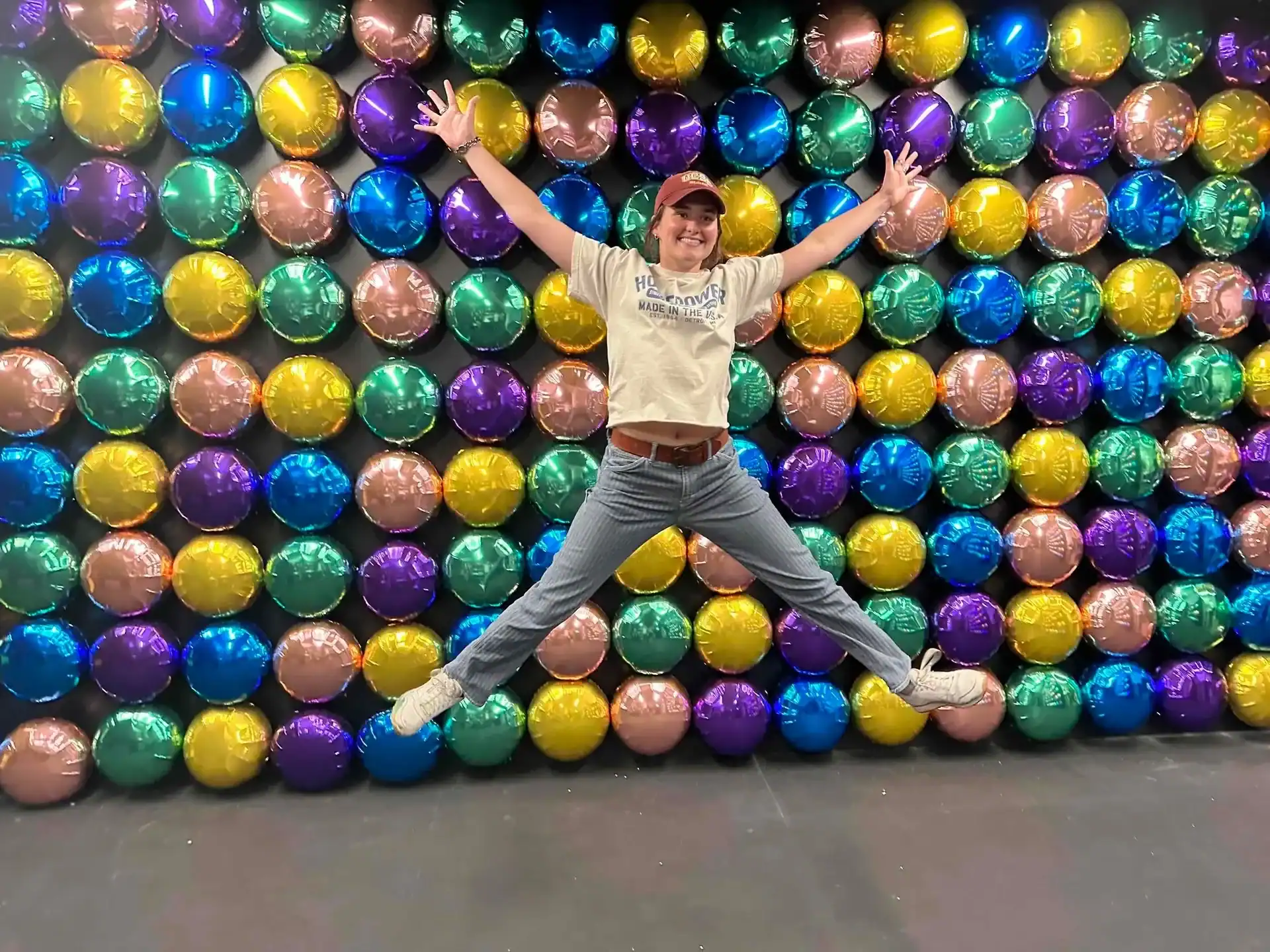 A person jumps with arms and legs spread in front of a colorful wall of metallic balloons arranged in a grid pattern, showcasing vibrant balloon installations perfect for any corporate event in Los Angeles.