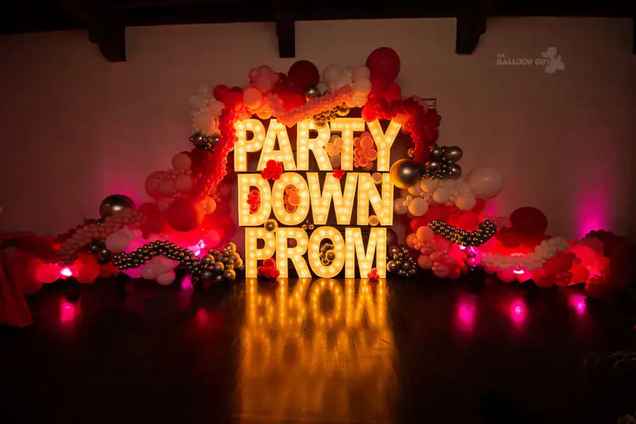 Large illuminated letters spelling "PARTY DOWN PROM" are surrounded by stunning balloon decor in clusters of red, pink, white, and silver against a dark background.