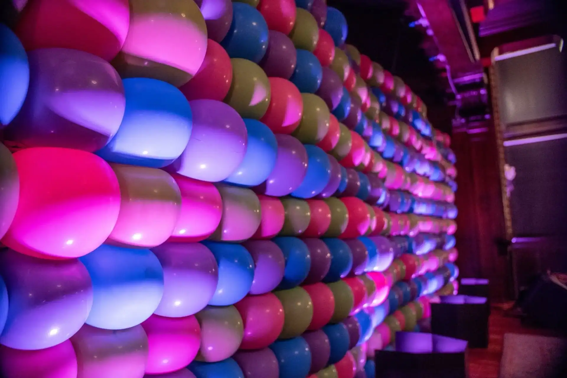 A wall covered with rows of colorful balloons—mainly pink, purple, and blue—glows under party lights in a dimly lit room, showcasing a stunning balloon installation by a Los Angeles balloon artist.