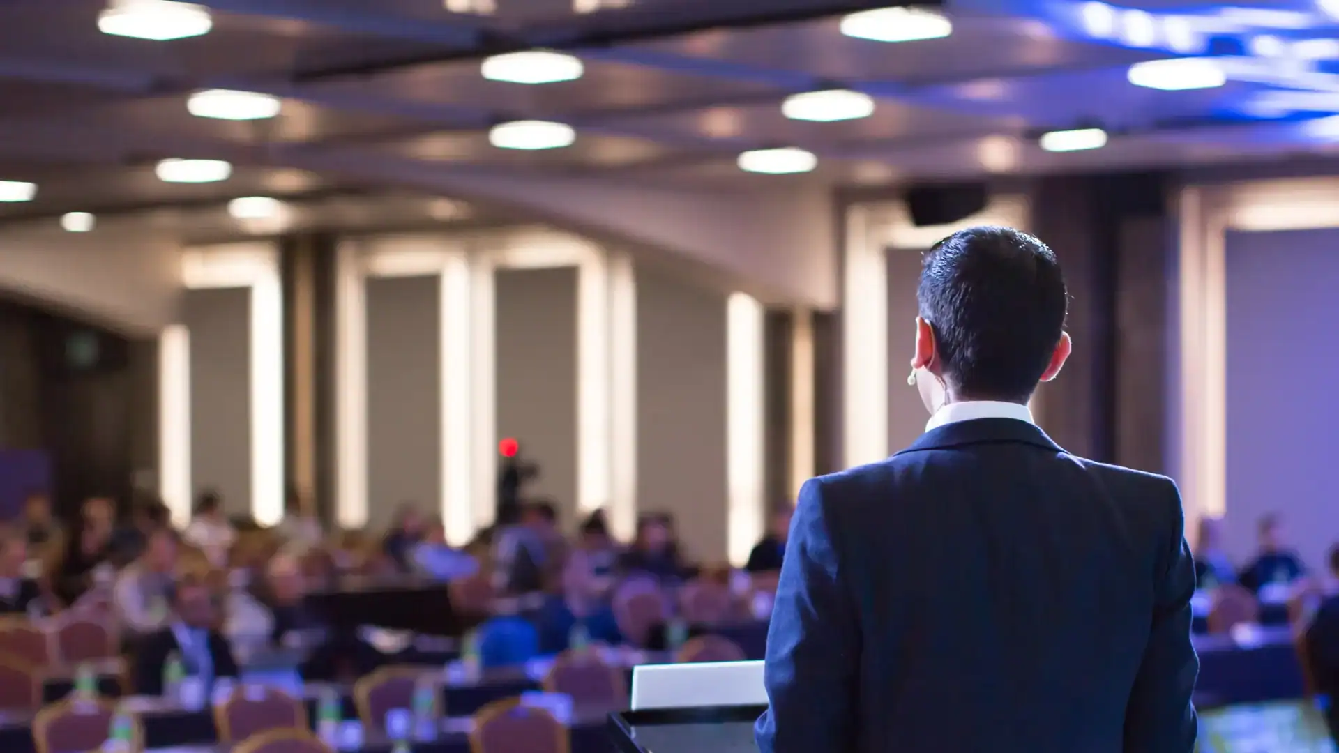 A man in a suit speaks at a podium in front of a seated audience in a brightly lit conference setting, accented by elegant balloon installations.