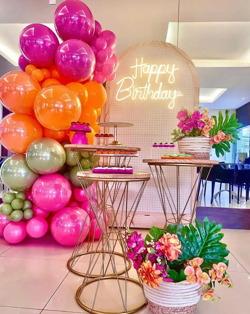 A birthday party setup featuring a neon "Happy Birthday" sign, vibrant balloon decor in pink and orange, gold stands with treats, and elegant flower arrangements.