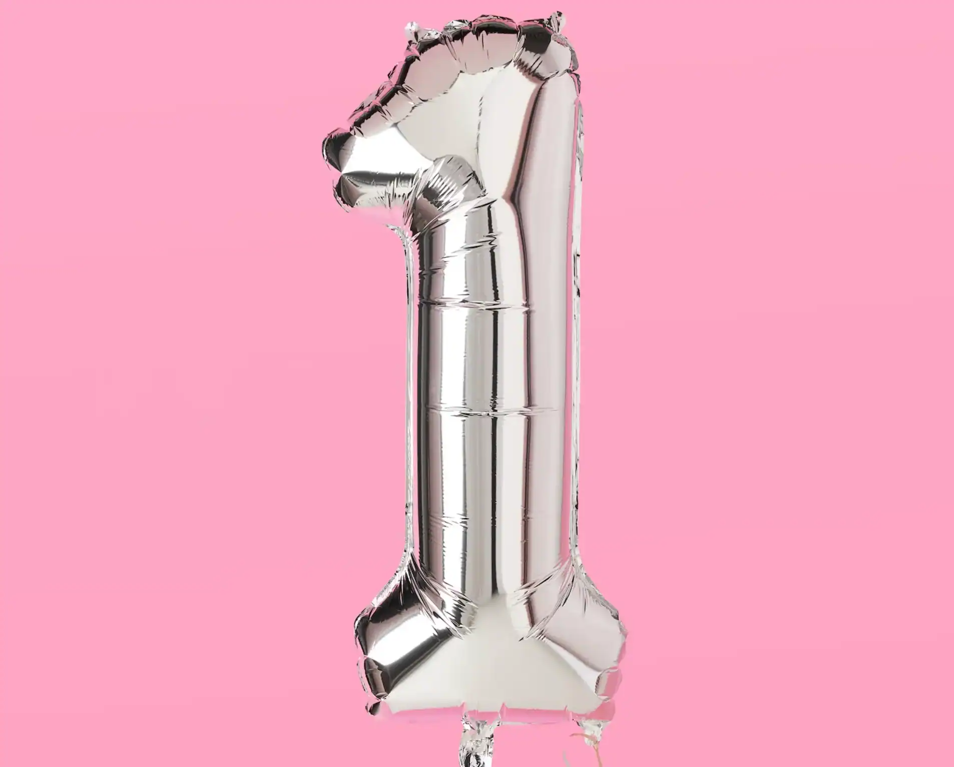 A metallic silver balloon shaped like the number one, crafted by a skilled balloon artist, stands out against a plain pink background.