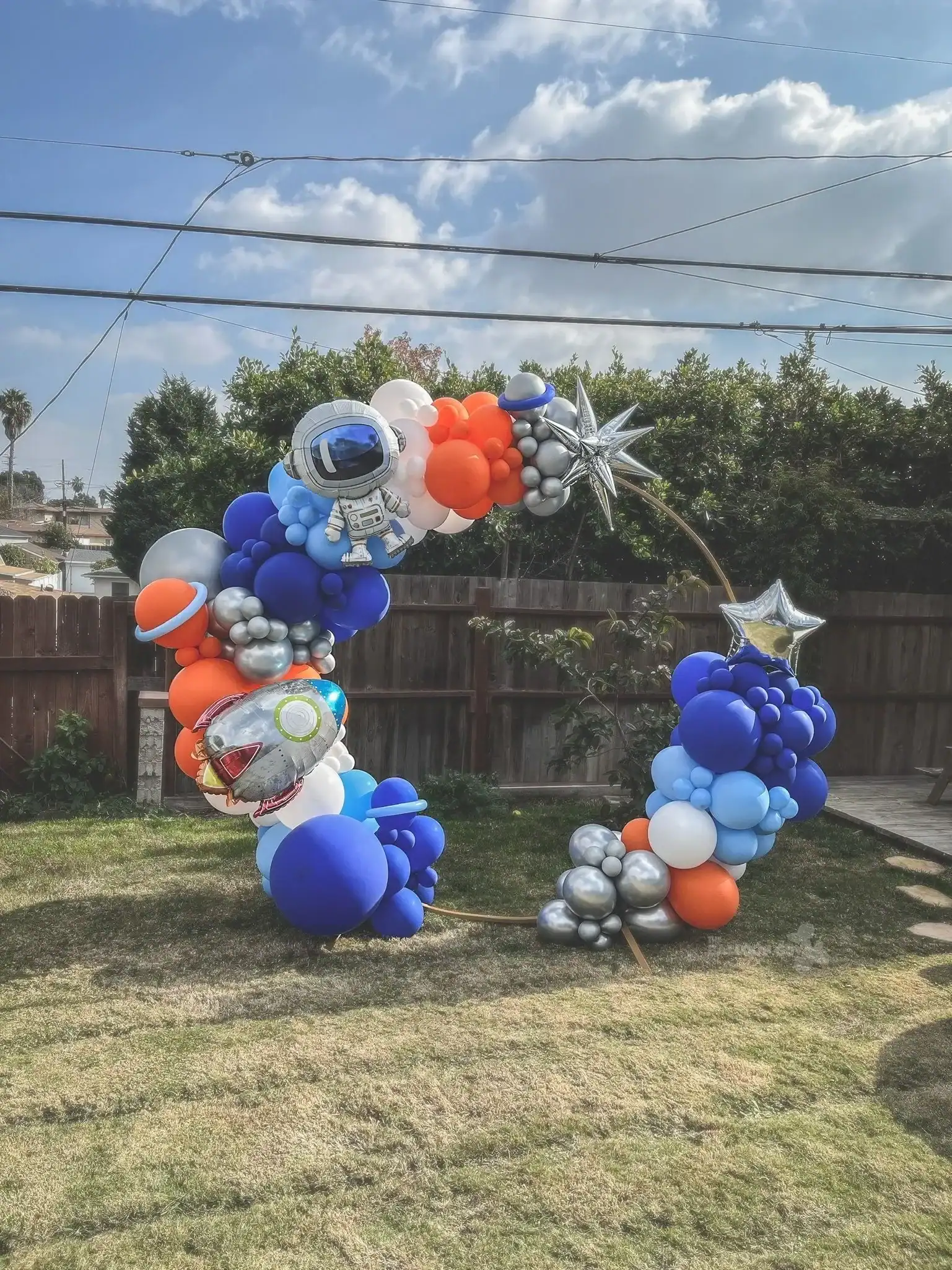 A balloon arch crafted by a talented balloon artist in Los Angeles features astronaut, rocket, and star balloons in blue, orange, silver, and white, standing on a grassy yard under a partly cloudy sky.