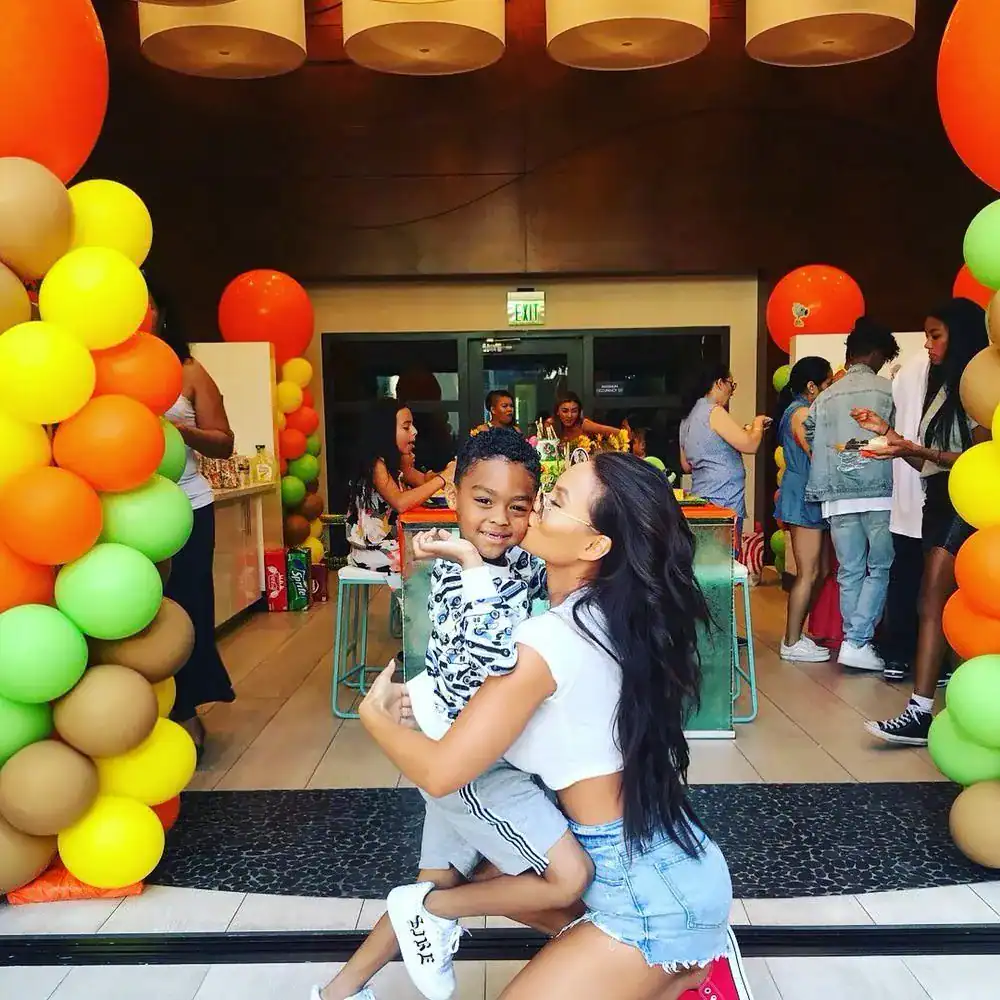 A woman hugs a smiling child at a colorful indoor party with balloons, delicious food, and several people in the background.