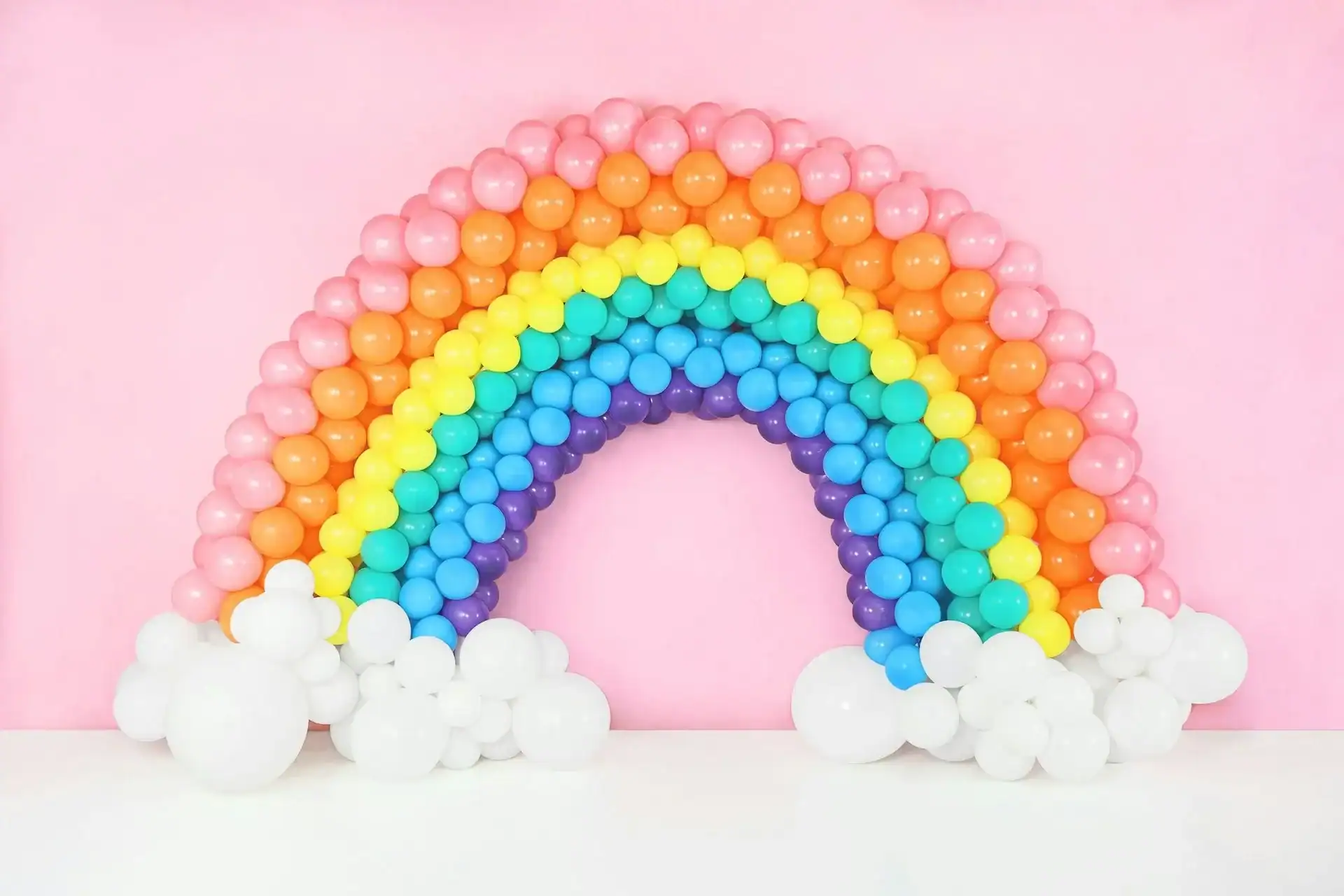 A rainbow made of colorful balloons is arranged against a pink wall, with white balloons at the base representing clouds—a stunning example of creative balloon decor.