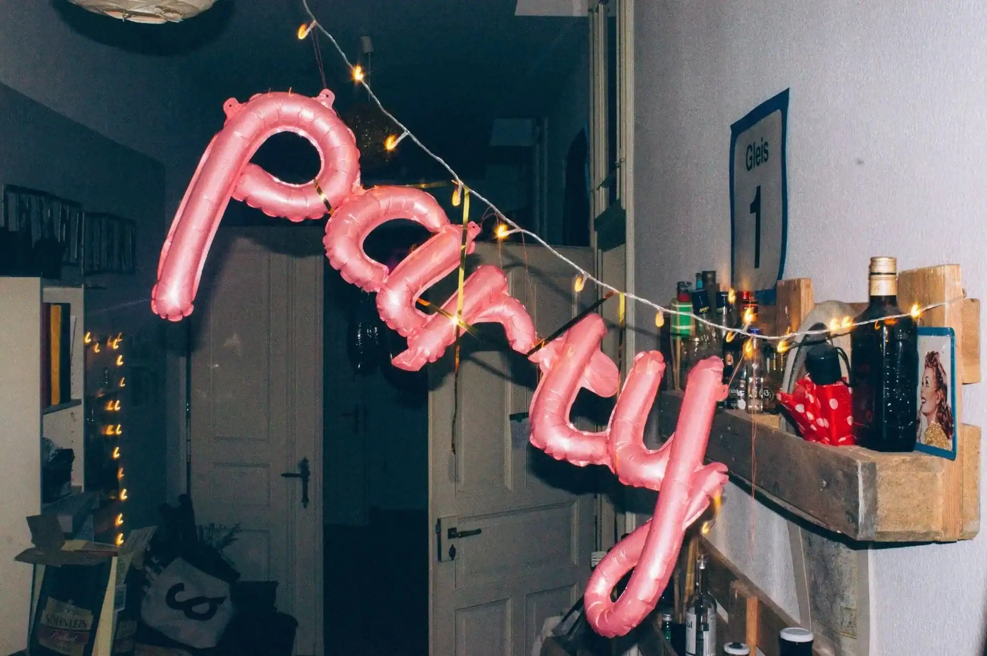 Pink foil balloon letters spelling "party," crafted by a skilled balloon artist, hang with string lights in a dimly lit Los Angeles hallway, with shelves of bottles and decor visible on the right—perfect for a stylish corporate event.