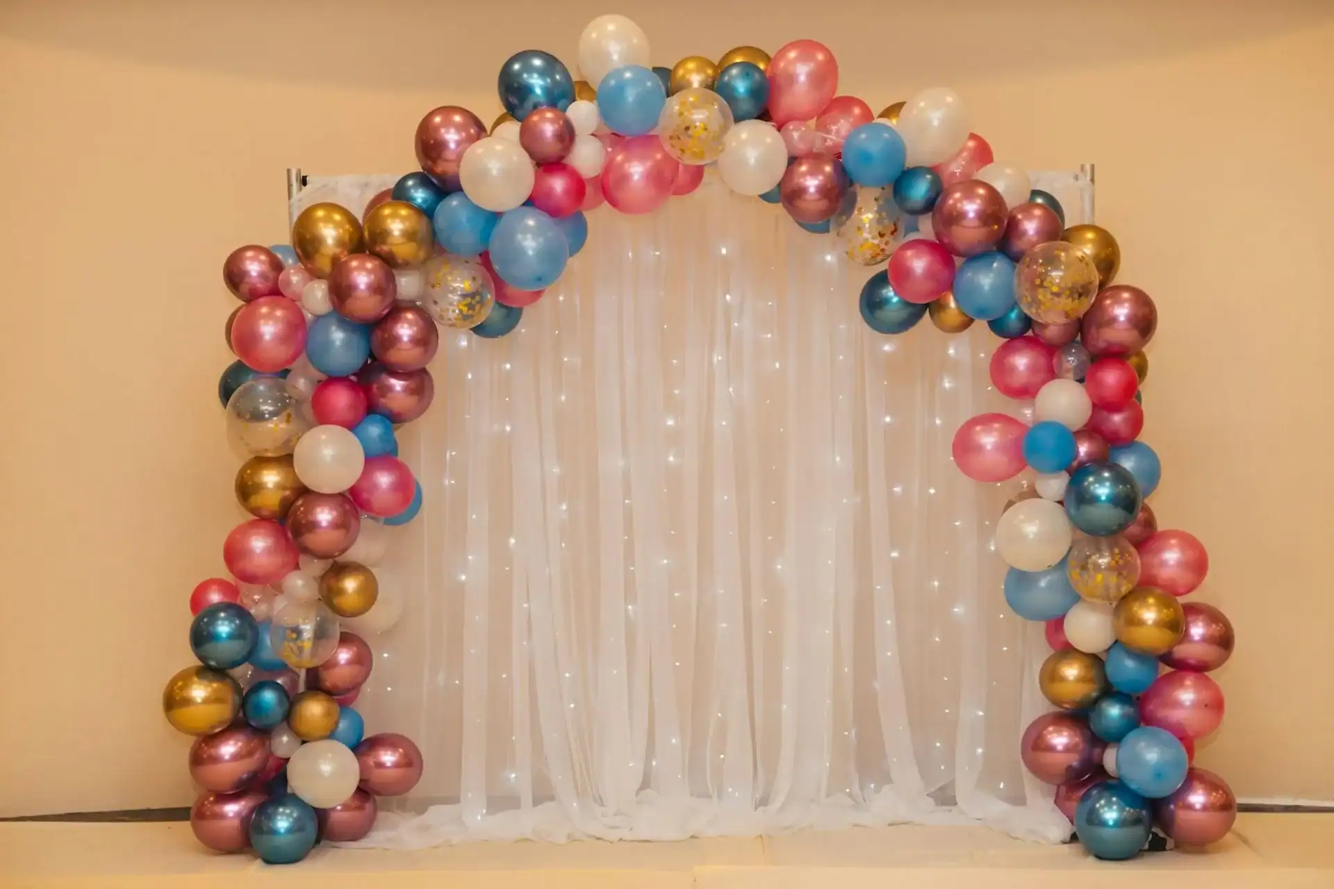 An arch of multicolored balloon decor, crafted by a skilled balloon artist, stands over white sheer curtains with string lights, set against a plain wall.