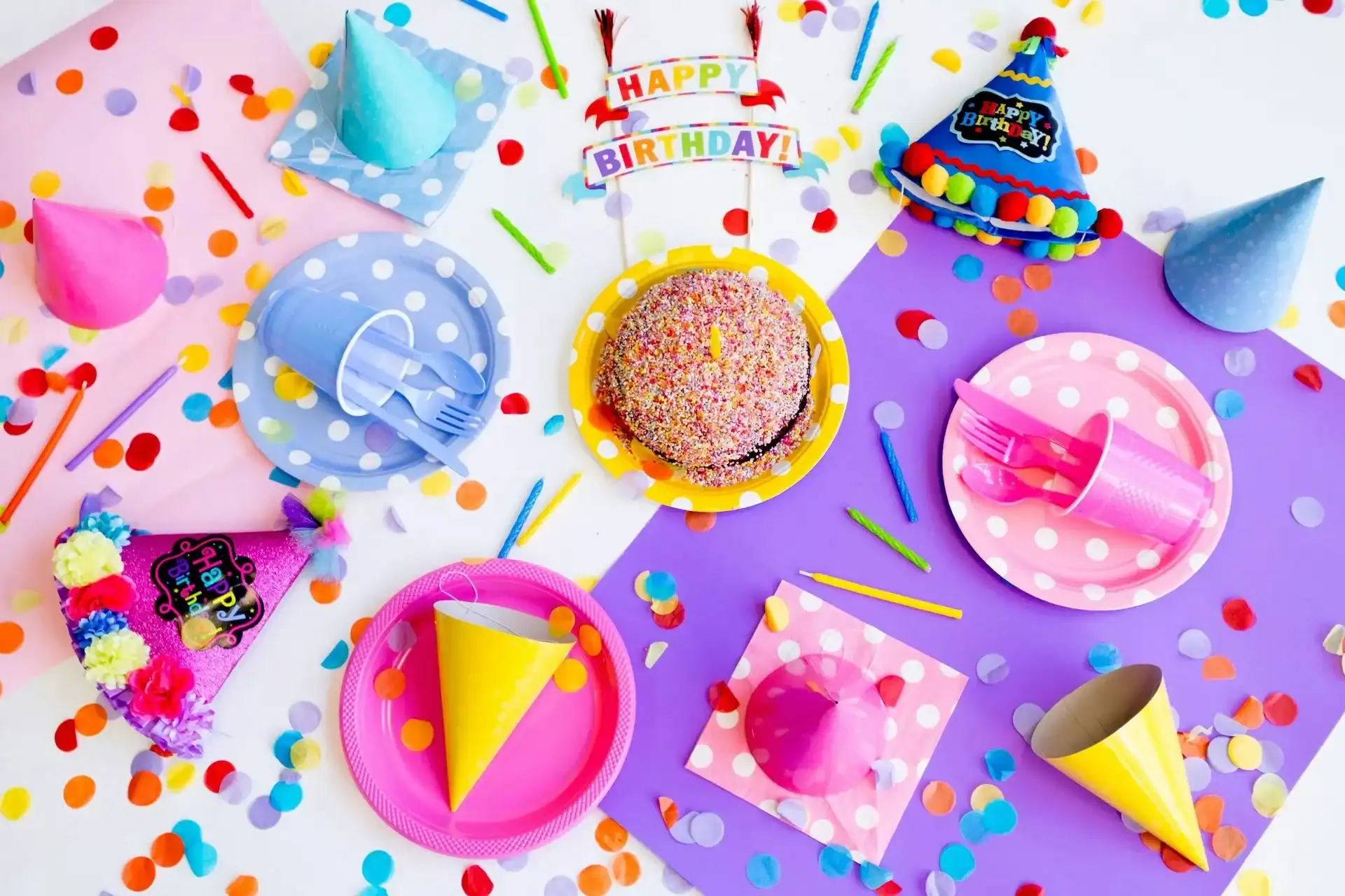 Colorful birthday party setup with a sprinkled cake, party hats, plates, utensils, napkins, confetti, and vibrant balloon decor on a white and purple background.