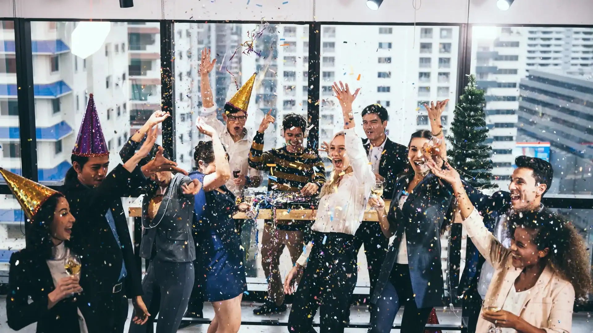 A group of people in business attire celebrate a corporate event indoors with confetti, party hats, and drinks, surrounded by balloon decor and standing in front of large windows overlooking the Los Angeles cityscape.