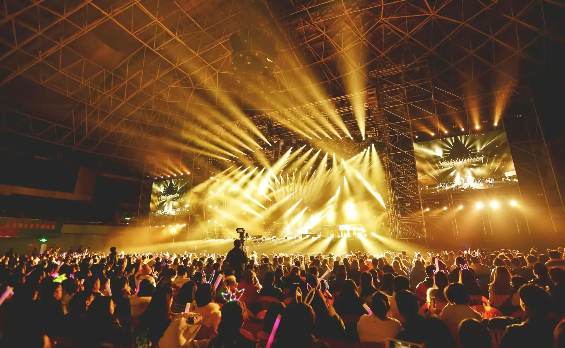 Large crowd seated at an indoor concert venue, facing a brightly lit stage with yellow spotlights, while vibrant balloon installations add a festive touch and a camera operator captures the excitement from the center aisle.