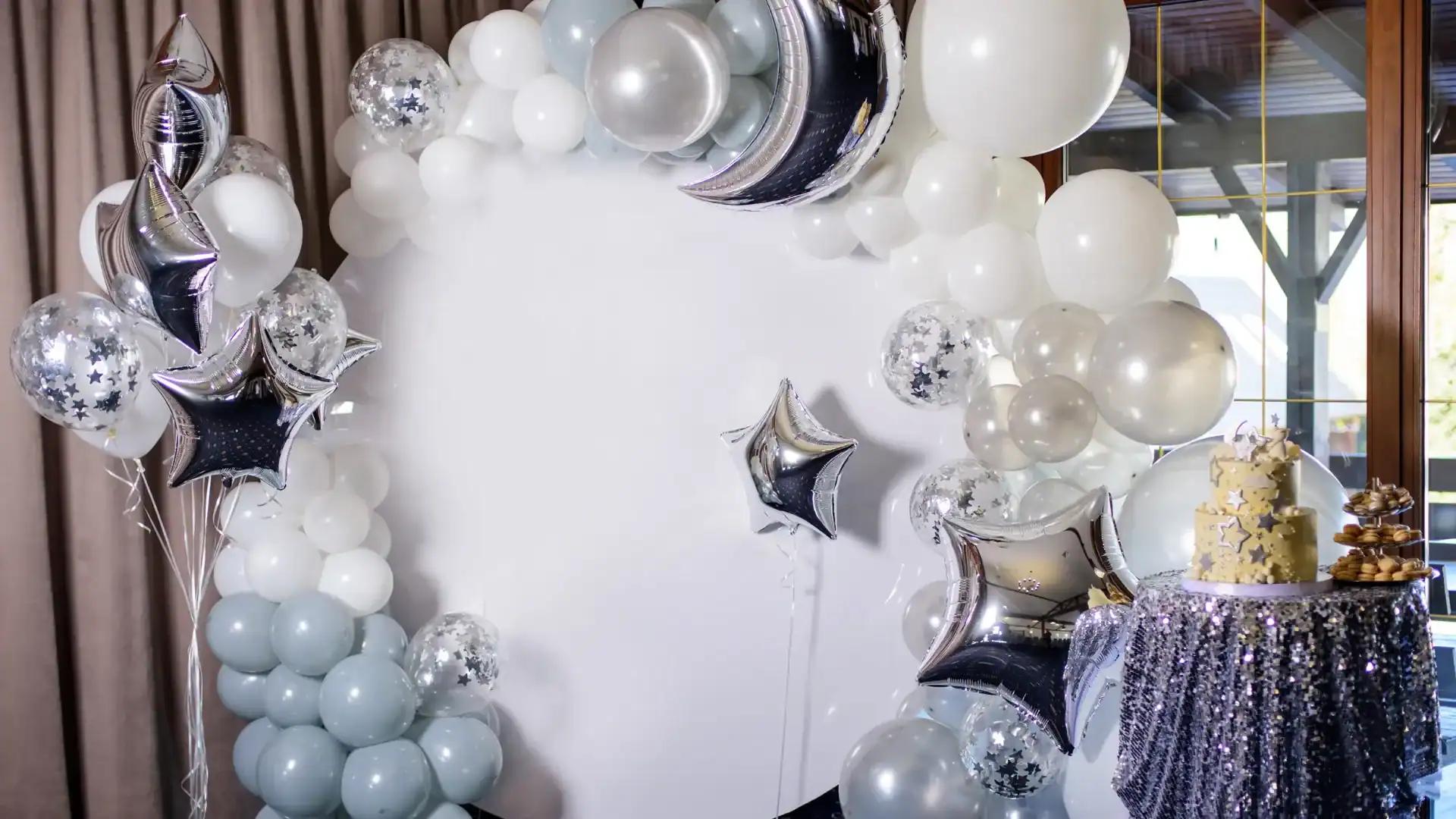 A stunning circular white balloon arch with silver and white balloons, star and moon foil balloons, and a decorated cake on a table to the right—perfect balloon decor for celebrations in Los Angeles.