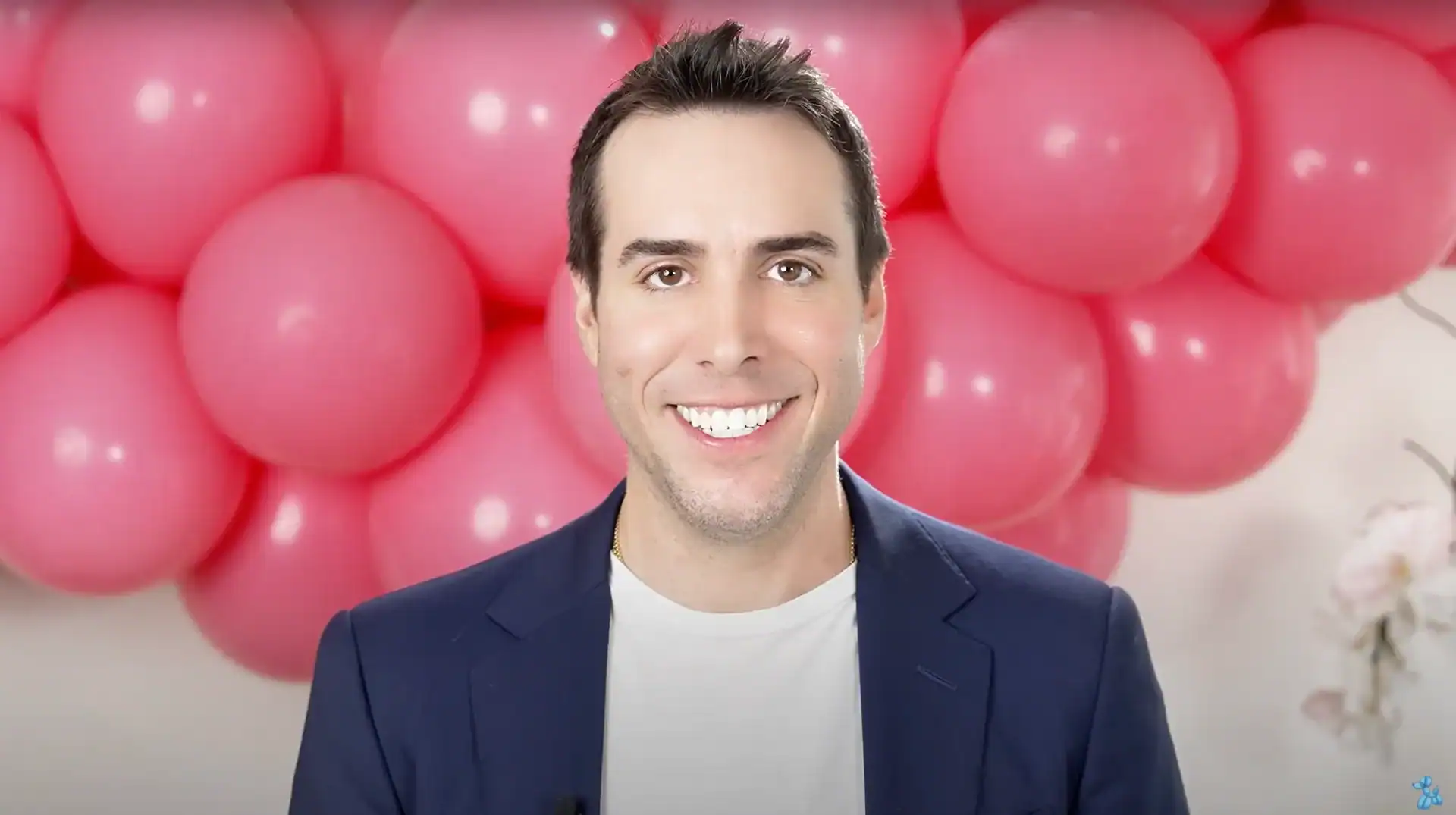 A man in a dark blazer and white shirt smiles at the camera, standing in front of a wall decorated with stunning balloon installations featuring large pink balloons.