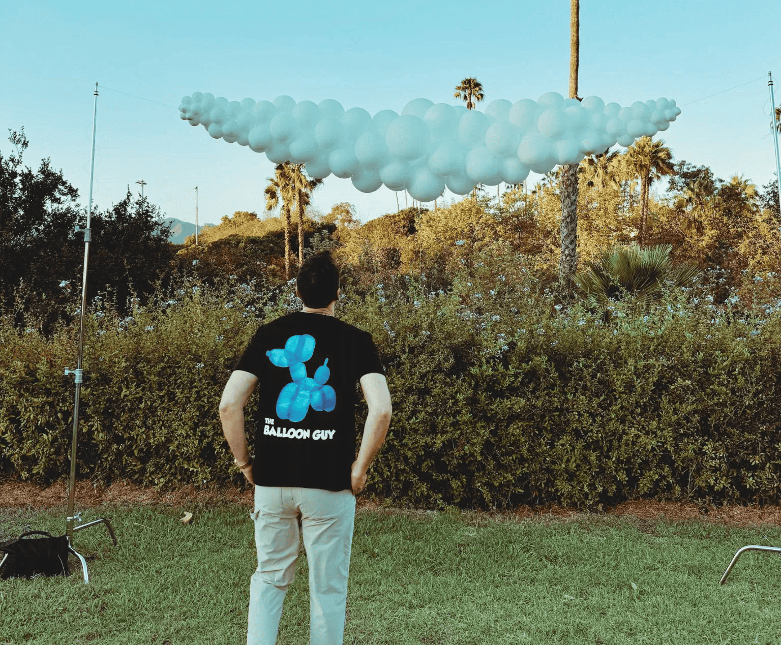 A person in a "Balloon Guy" shirt stands on grass at a Los Angeles corporate event, admiring a large cluster of white balloon decor suspended in the air outdoors.