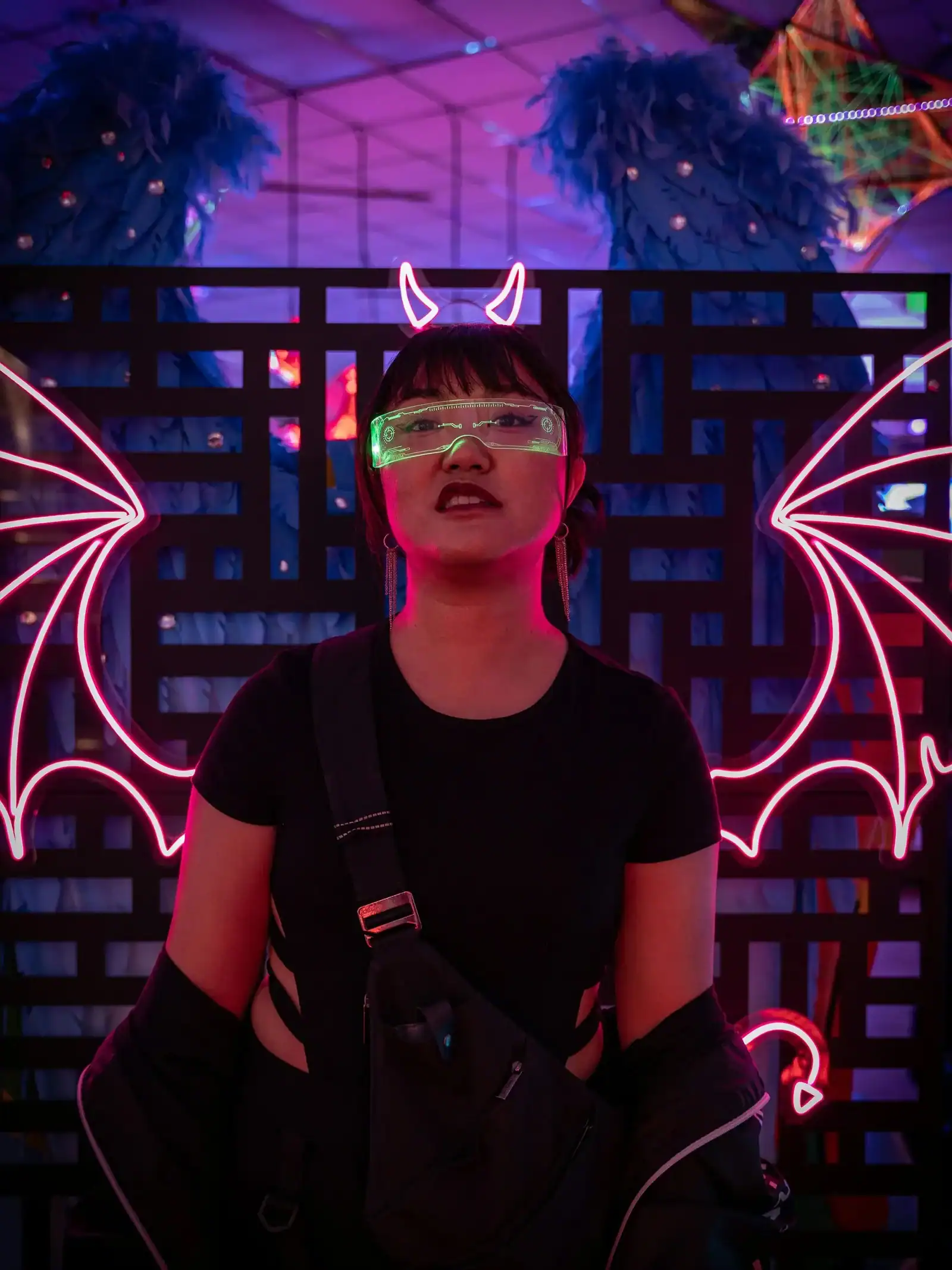 Person stands in front of neon lights shaped like bat wings and devil horns, wearing reflective glasses and dark clothing, at a colorful indoor event in Los Angeles.