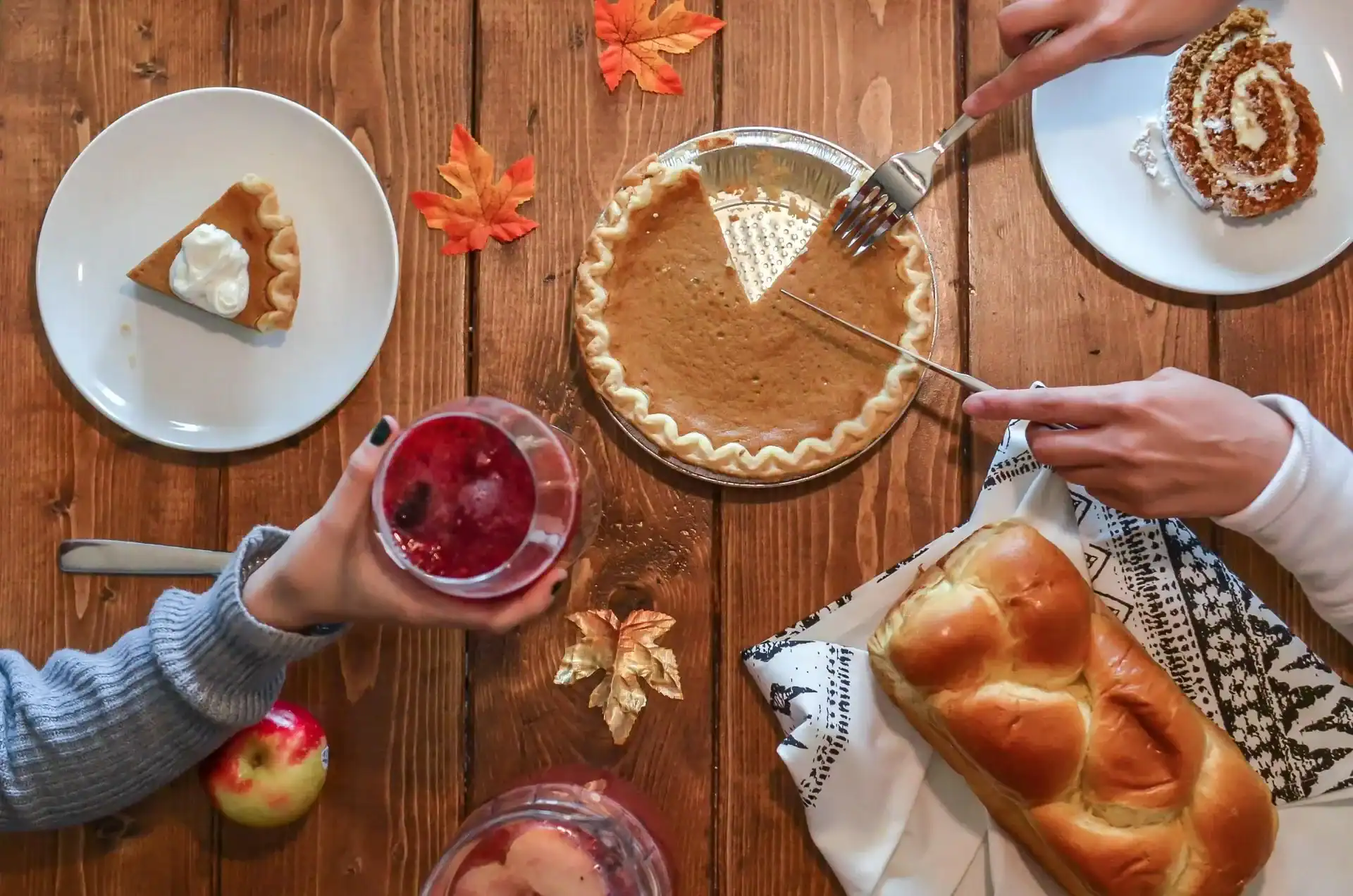 A table set with pumpkin pie, braided bread, a slice of pie on a plate, an apple, and a glass of red drink. Two hands are serving pie. Autumn leaves and elegant balloon decor create a festive atmosphere.