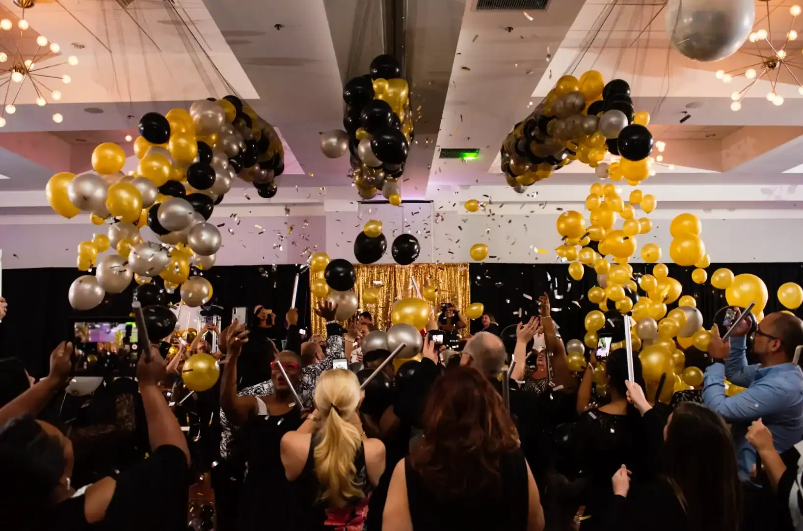A crowd in formal attire celebrates indoors in Los Angeles as black, gold, and silver balloons fall from the ceiling, with confetti and raised phones capturing the festive atmosphere.