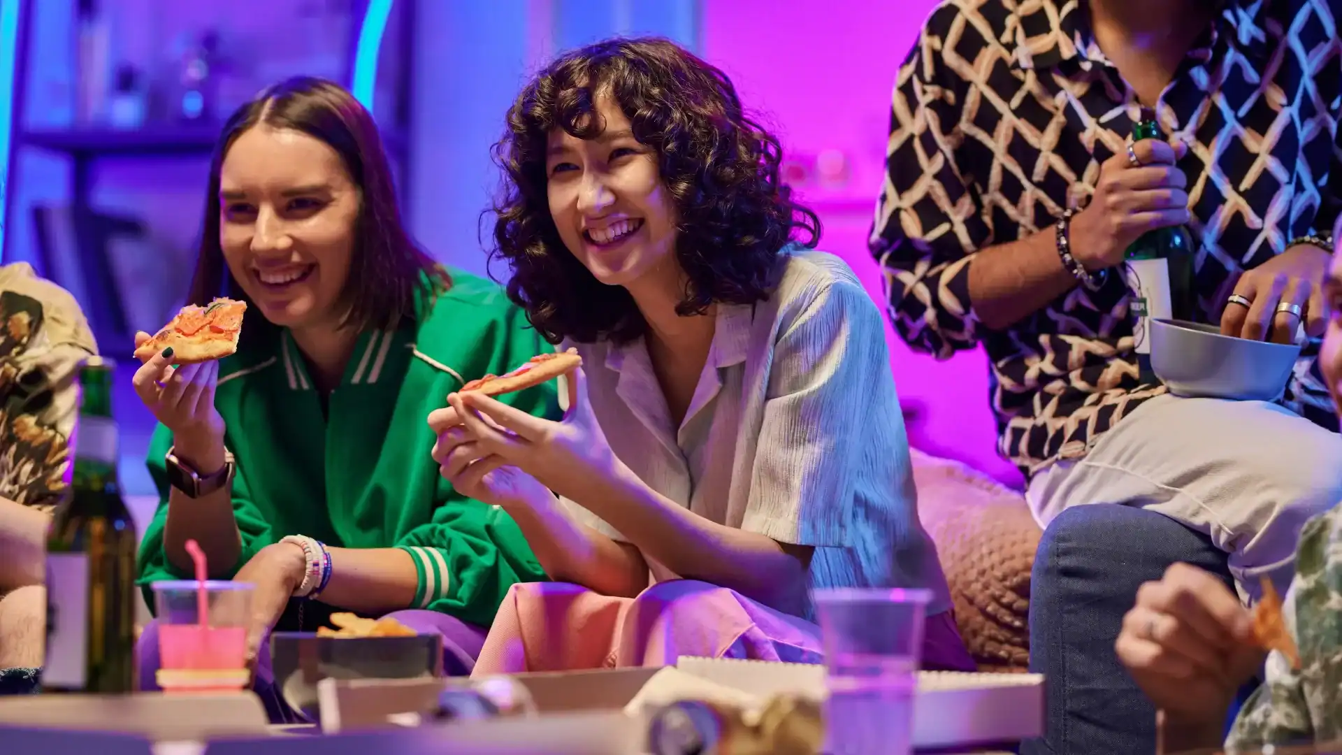 Three people sit together indoors at a corporate event, eating and smiling, with colorful lighting and festive balloon decor in the background and food and drinks on the table in front of them.
