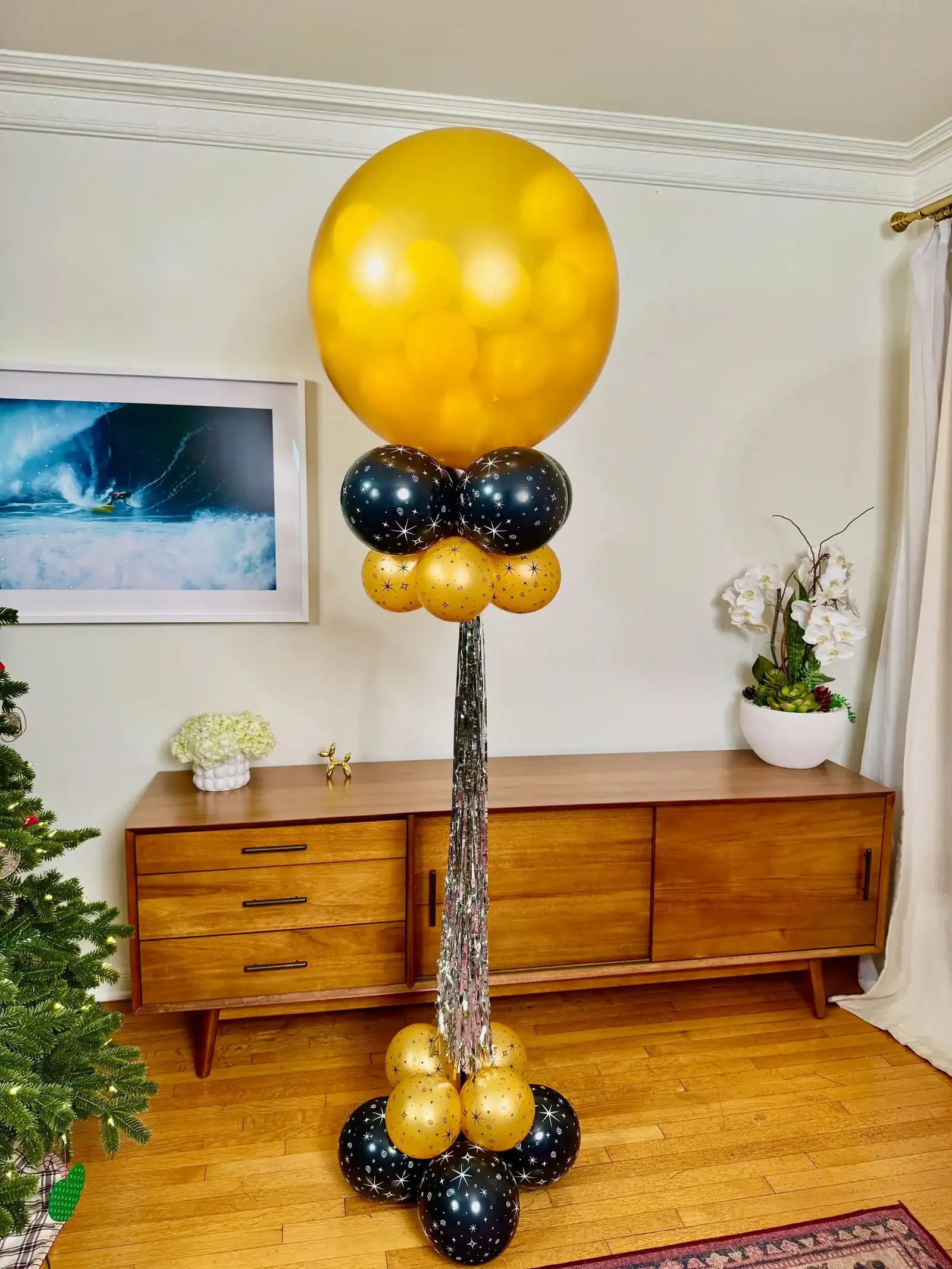 A tall balloon arrangement by a skilled balloon artist features a large gold balloon on top, smaller black and gold balloons at the base, and silver streamers, standing in a Los Angeles living room next to a wooden credenza.