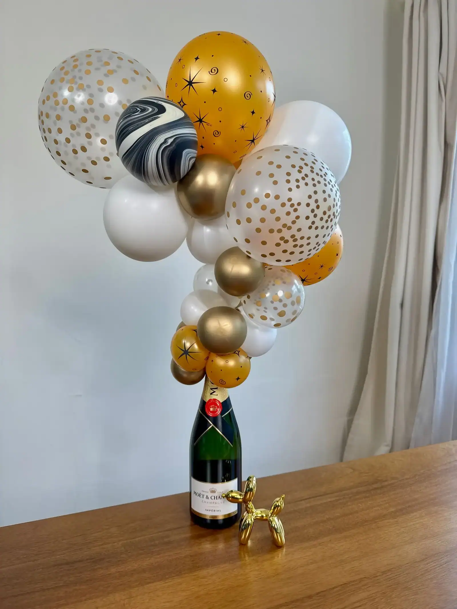A champagne bottle is topped with a decorative arrangement of gold, white, and patterned balloons, creating elegant balloon decor on a wooden table in Los Angeles, with a small gold balloon dog sculpture nearby.