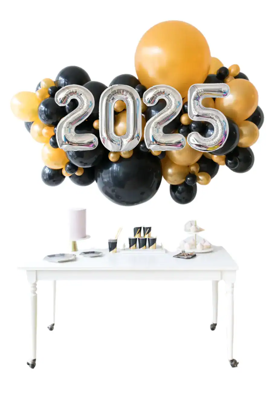 A white table with party supplies is set in front of striking balloon decor, featuring a black and gold balloon arrangement and large silver balloons spelling "2025.