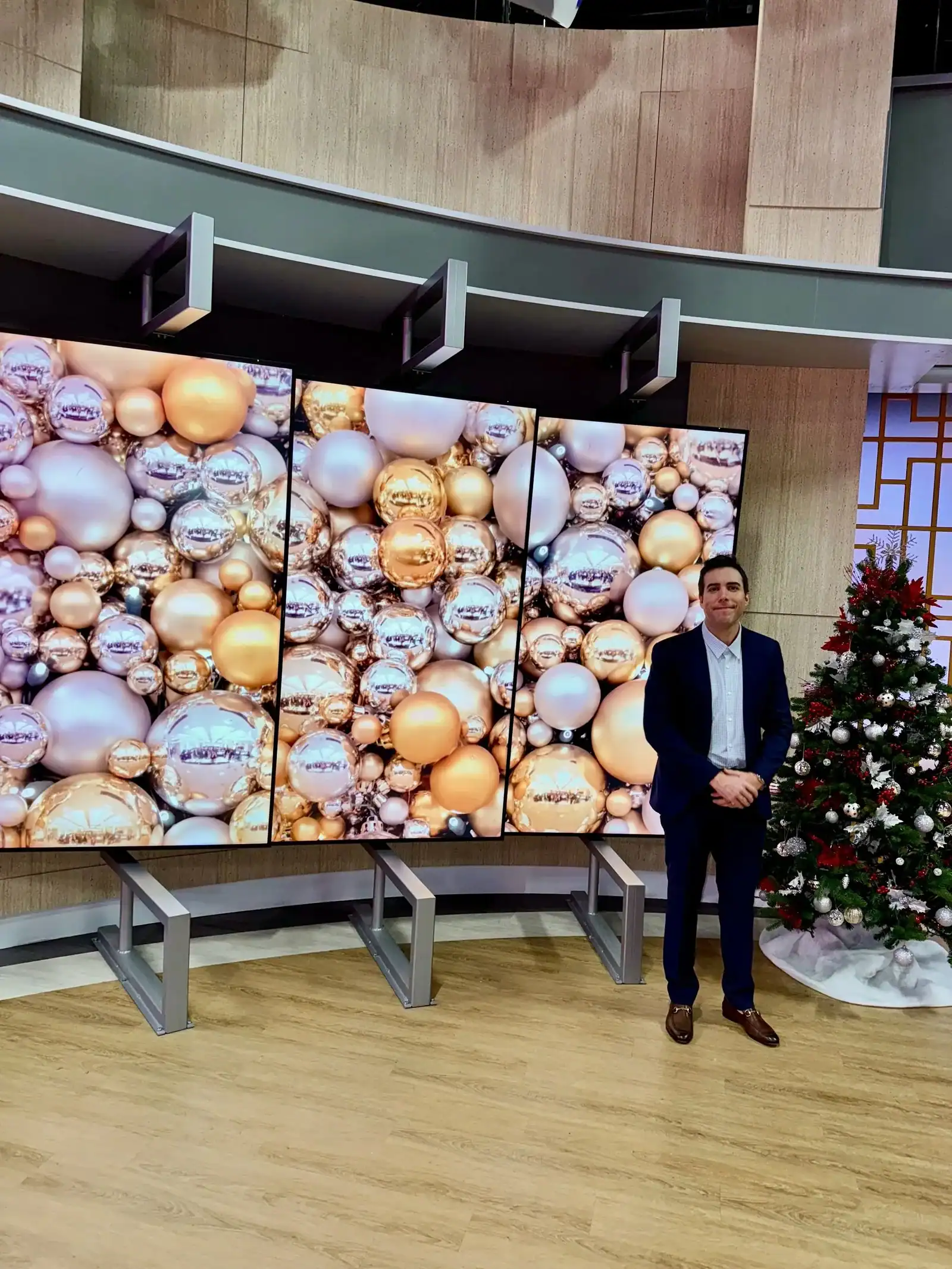 A man in a dark suit stands next to a decorated Christmas tree and large display screens showing a close-up of gold and silver holiday ornaments at a corporate event.