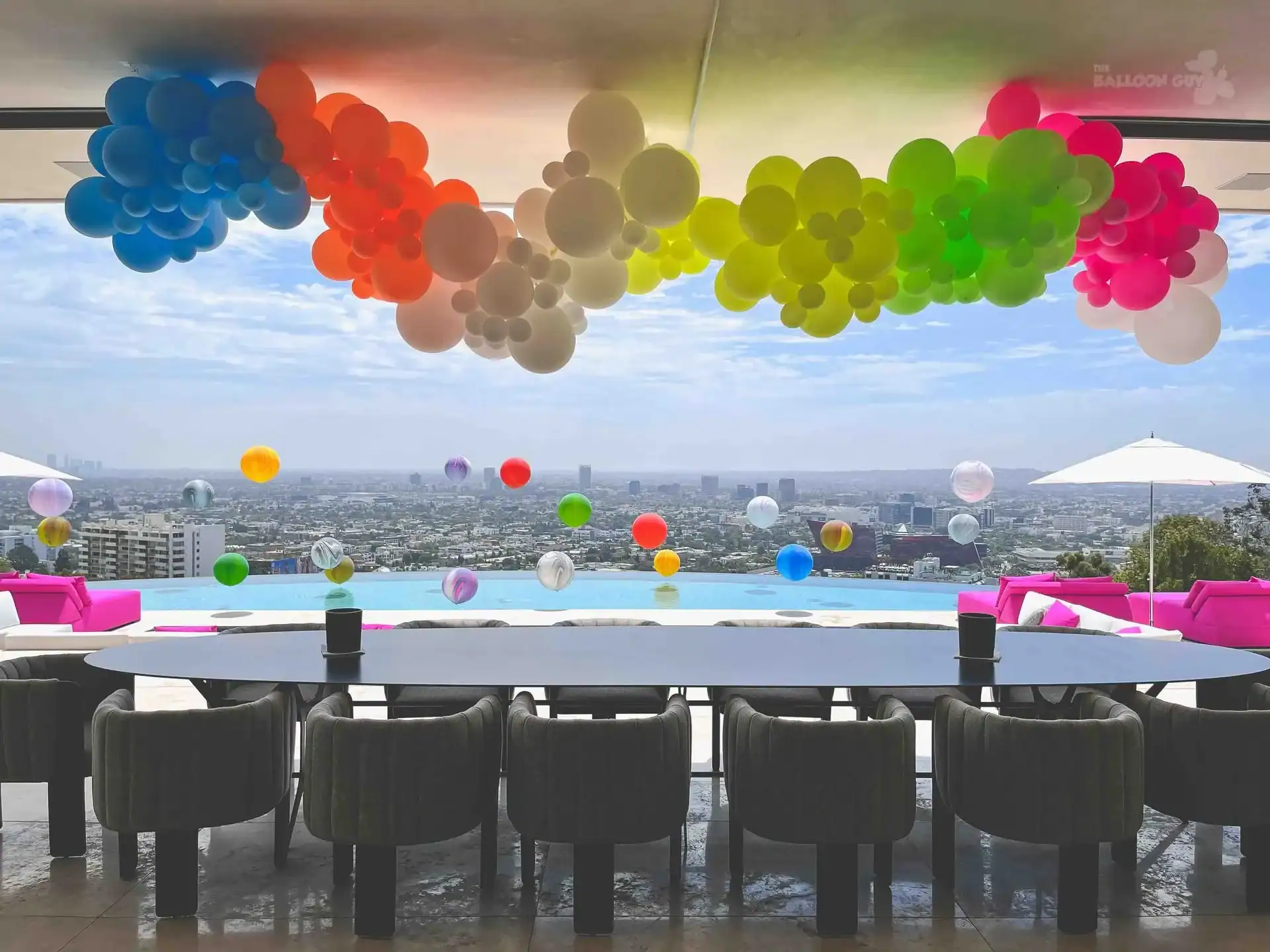 A long table with chairs overlooks a pool and cityscape, enhanced by vibrant balloon installations arranged above and across the pool area.