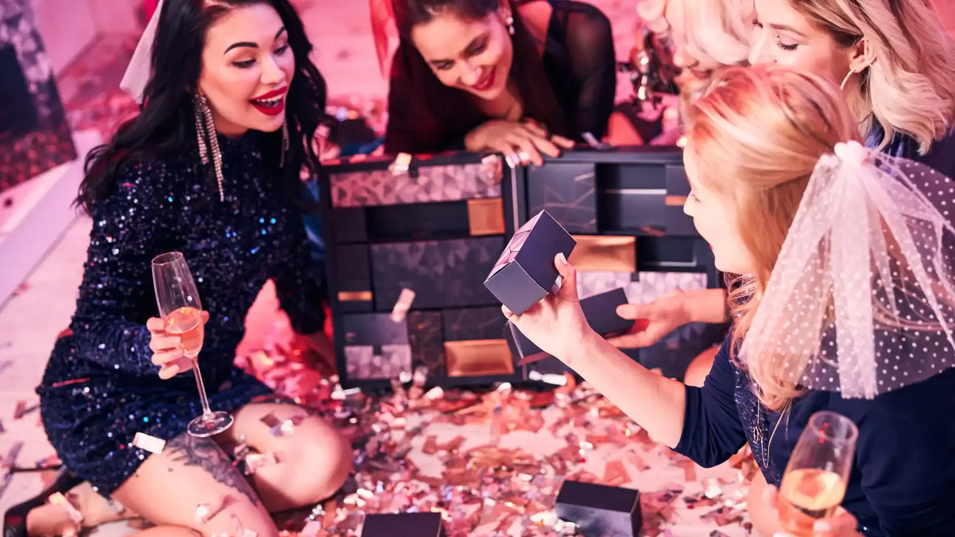 Four women in festive attire sit on the floor in Los Angeles, surrounded by confetti, colorful gift boxes, and vibrant balloon decor, with one woman holding up a small box.