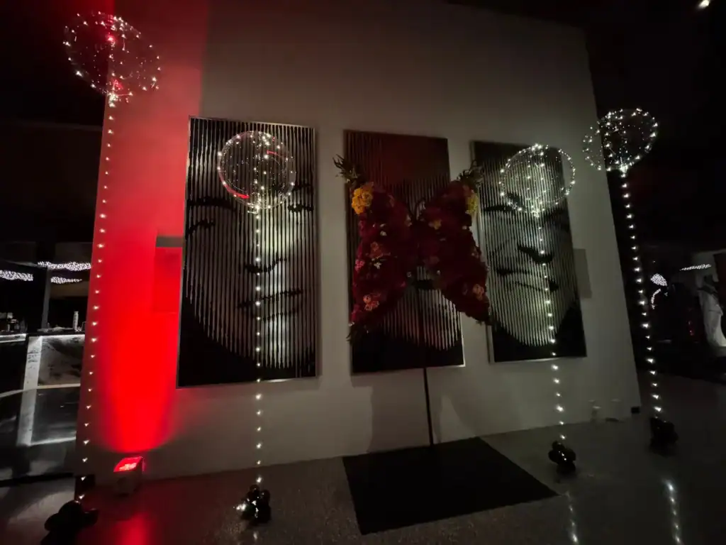 Three large grayscale portrait panels are displayed on a wall at a Los Angeles corporate event, with a central floral butterfly arrangement and illuminated string lights creating a vertical dotted pattern.