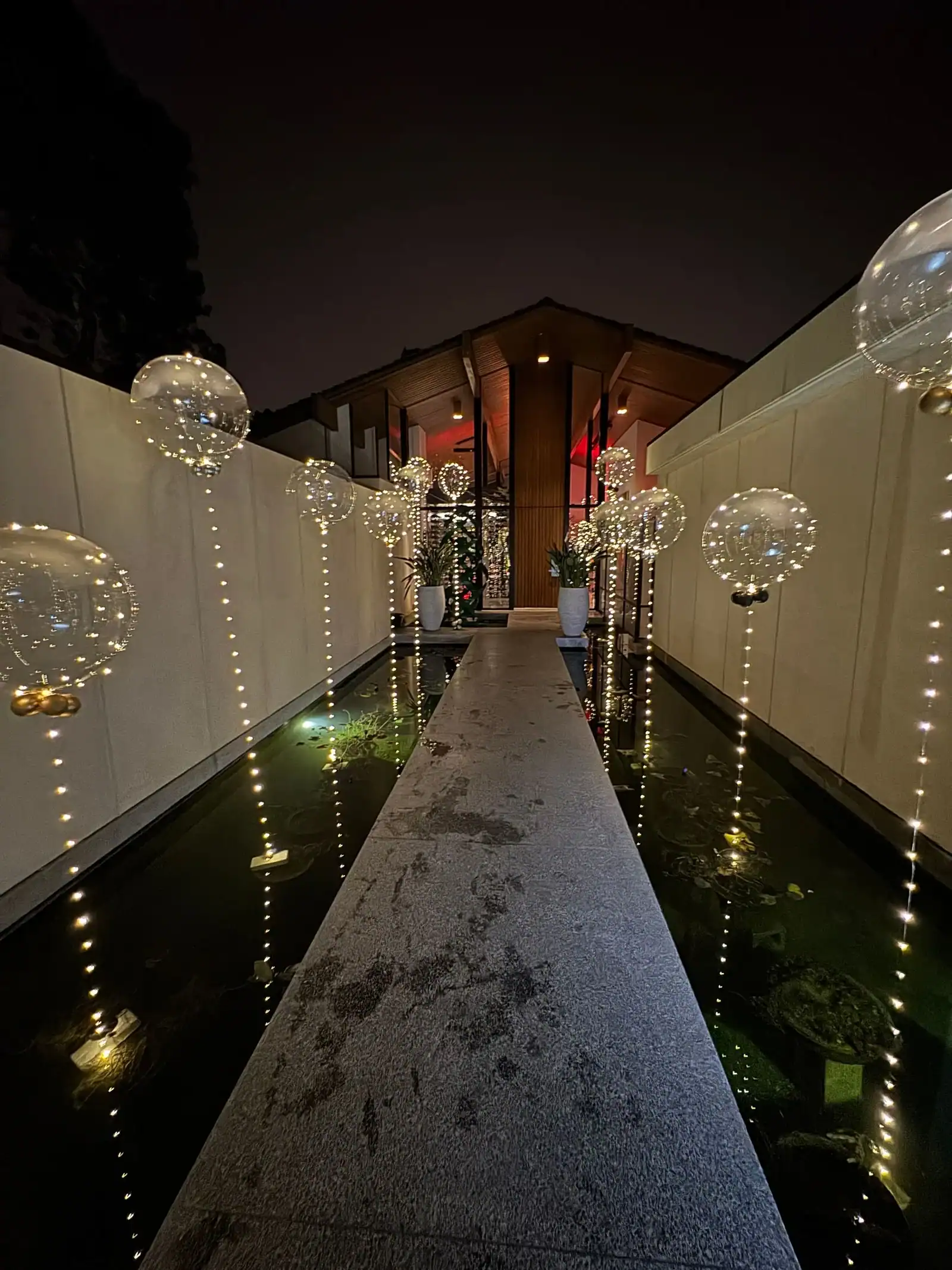 A nighttime walkway over water, lined with illuminated glass globes and string lights, leads to a modern building with a high, pointed roof—perfect for a corporate event enhanced by elegant balloon decor.