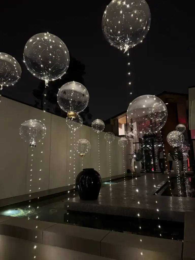 Clear balloons filled with string lights float above a modern outdoor water feature at night, creating a decorative and illuminated scene that showcases the creative flair of a talented Los Angeles balloon artist.