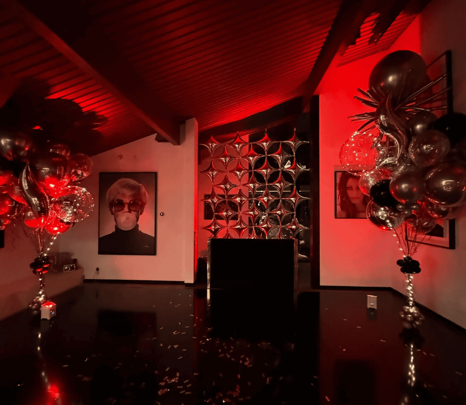 A dimly lit Los Angeles room with red lighting, striking balloon installations on either side, framed portraits on the walls, and a reflective black floor.