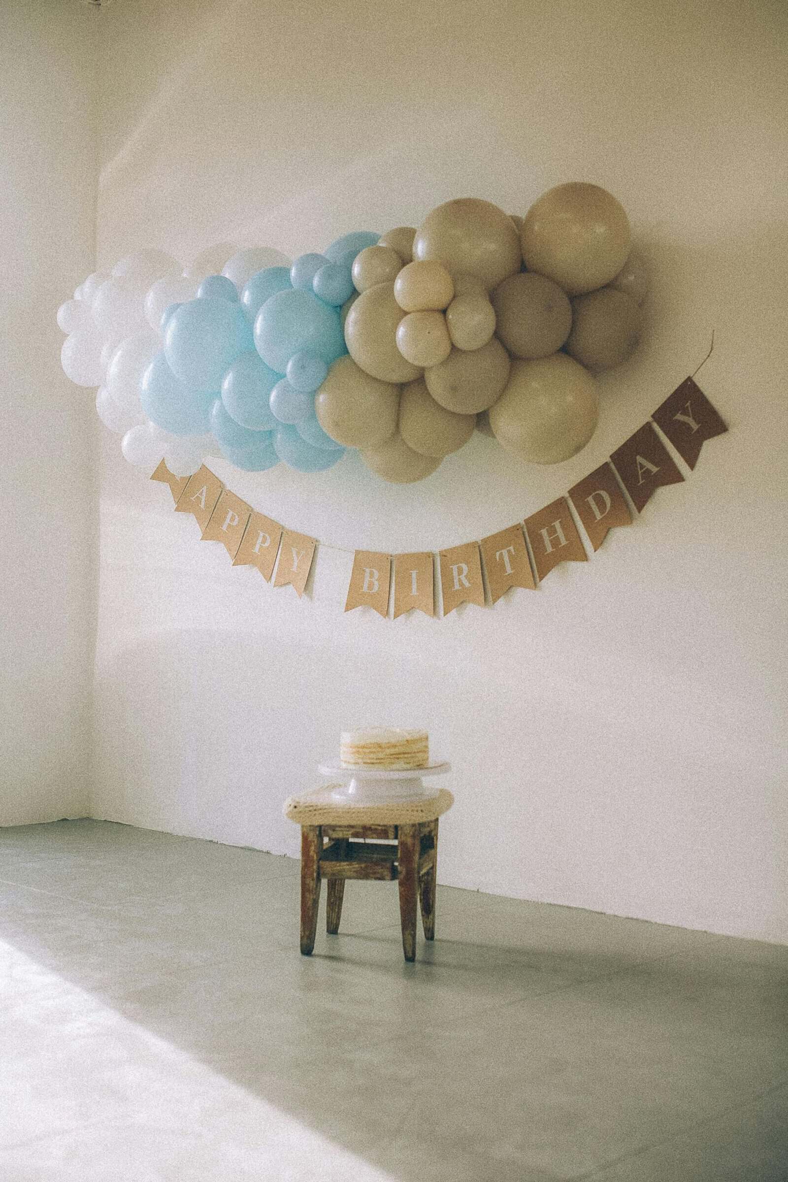 A birthday setup with a small cake on a stool, beige and blue balloon decor on the wall, and a banner reading "Happy Birthday.